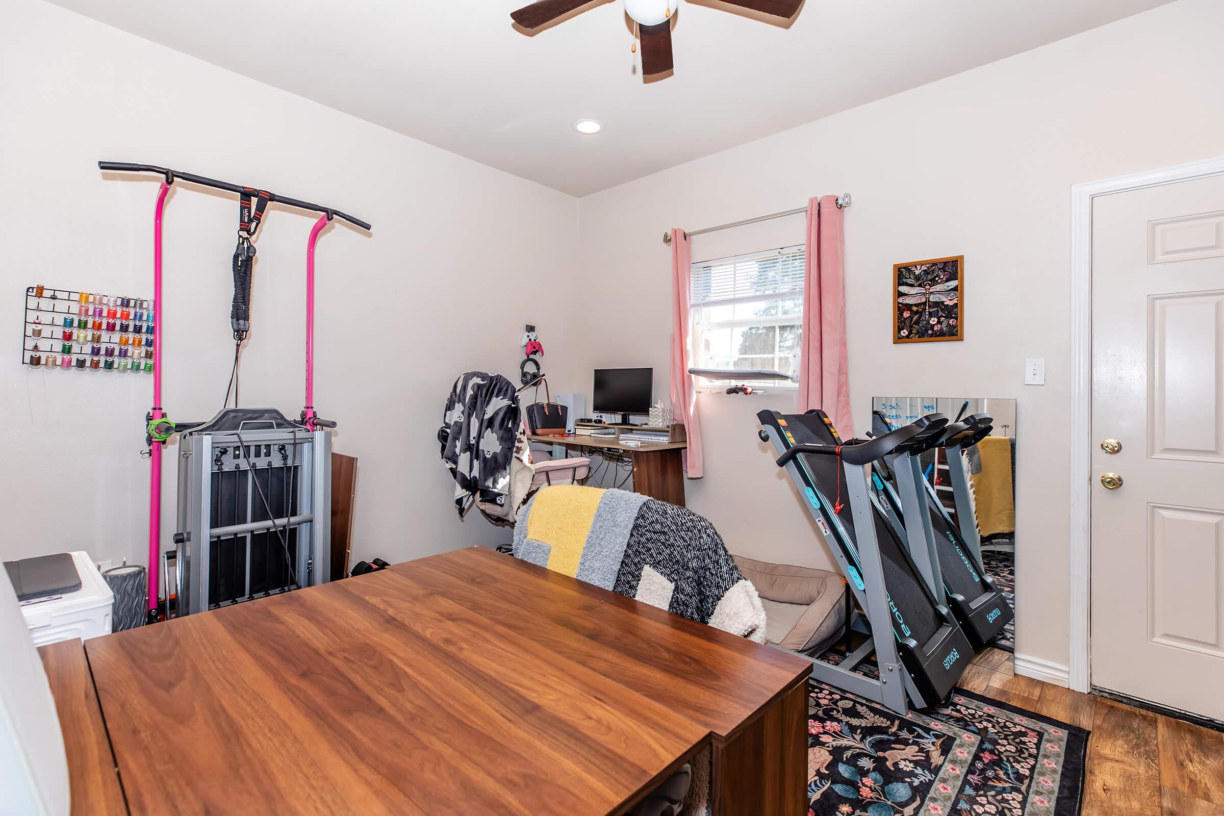 A home office featuring a wooden desk, a computer setup, and a wall-mounted exercise pull-up bar. There is a treadmill, an exercise mat, and various decor items, including a framed picture. The room has a warm atmosphere with light-colored walls and wooden flooring.