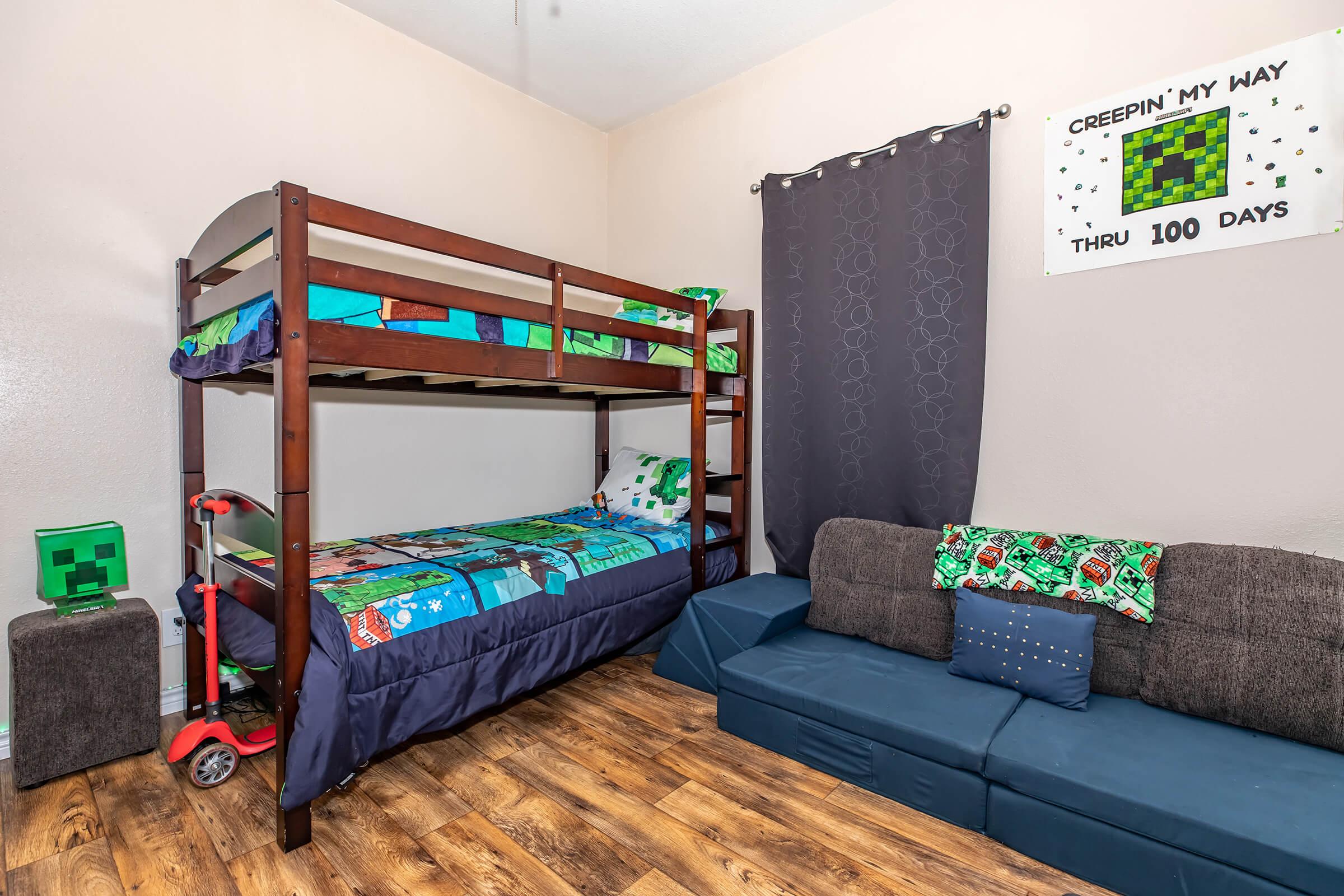 A children's bedroom featuring a wooden bunk bed with themed bedding, a cozy couch, and a green block decoration. The wall decor includes a poster with the phrase "CREEPIN' MY WAY THRU 100 DAYS." The room has wood-like flooring and a dark curtain, creating a playful atmosphere for kids.