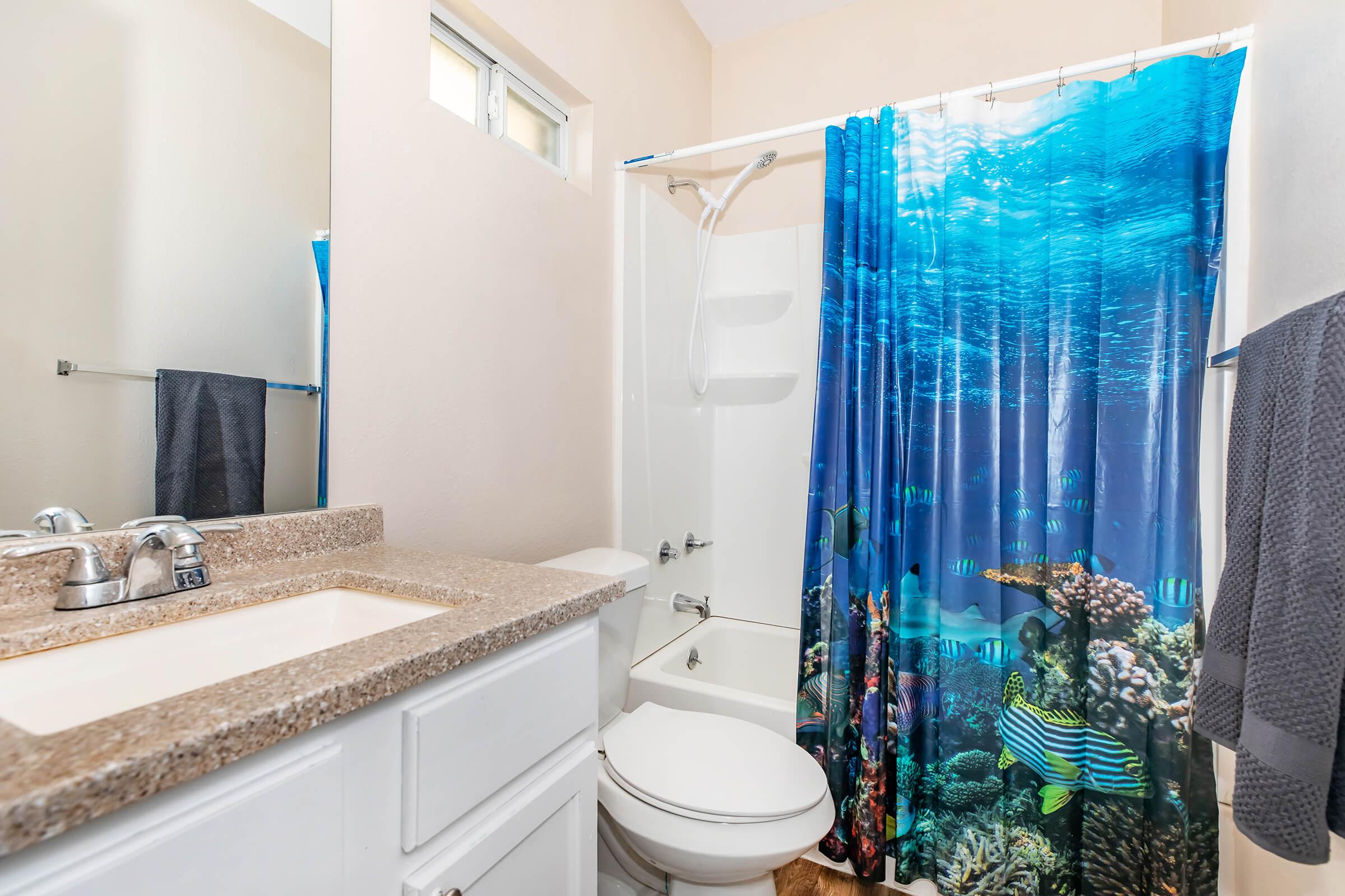 A well-lit bathroom featuring a shower with a colorful underwater-themed curtain, a beige wall, a countertop with a sink, and a toilet. There's a towel rack with a dark gray towel and a mirror above the sink. The overall decor is clean and modern.
