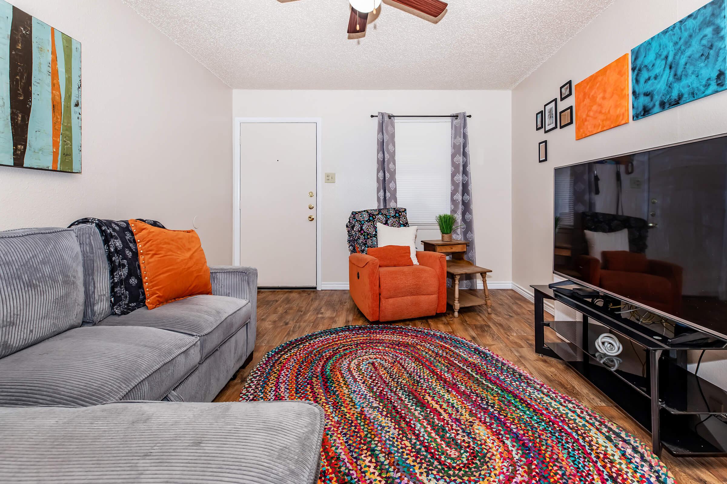 Cozy living room featuring a gray couch with orange pillows, a bright woven area rug, an orange armchair, a wooden side table, and a large TV. The walls are adorned with colorful art pieces, and there's a window with light curtains letting in natural light. A simple front door is visible.