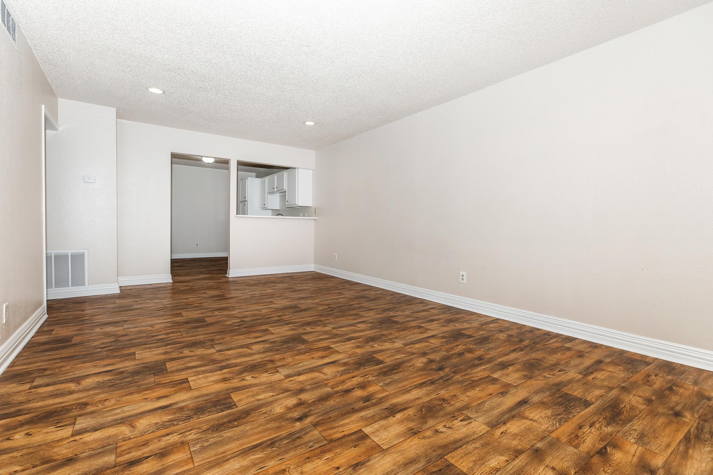 A spacious, empty room with light brown wood-like flooring and beige walls. The room features recessed lighting and a view of an adjacent area with a kitchen and additional space. The overall aesthetic is modern and bright, ideal for various interior design options.