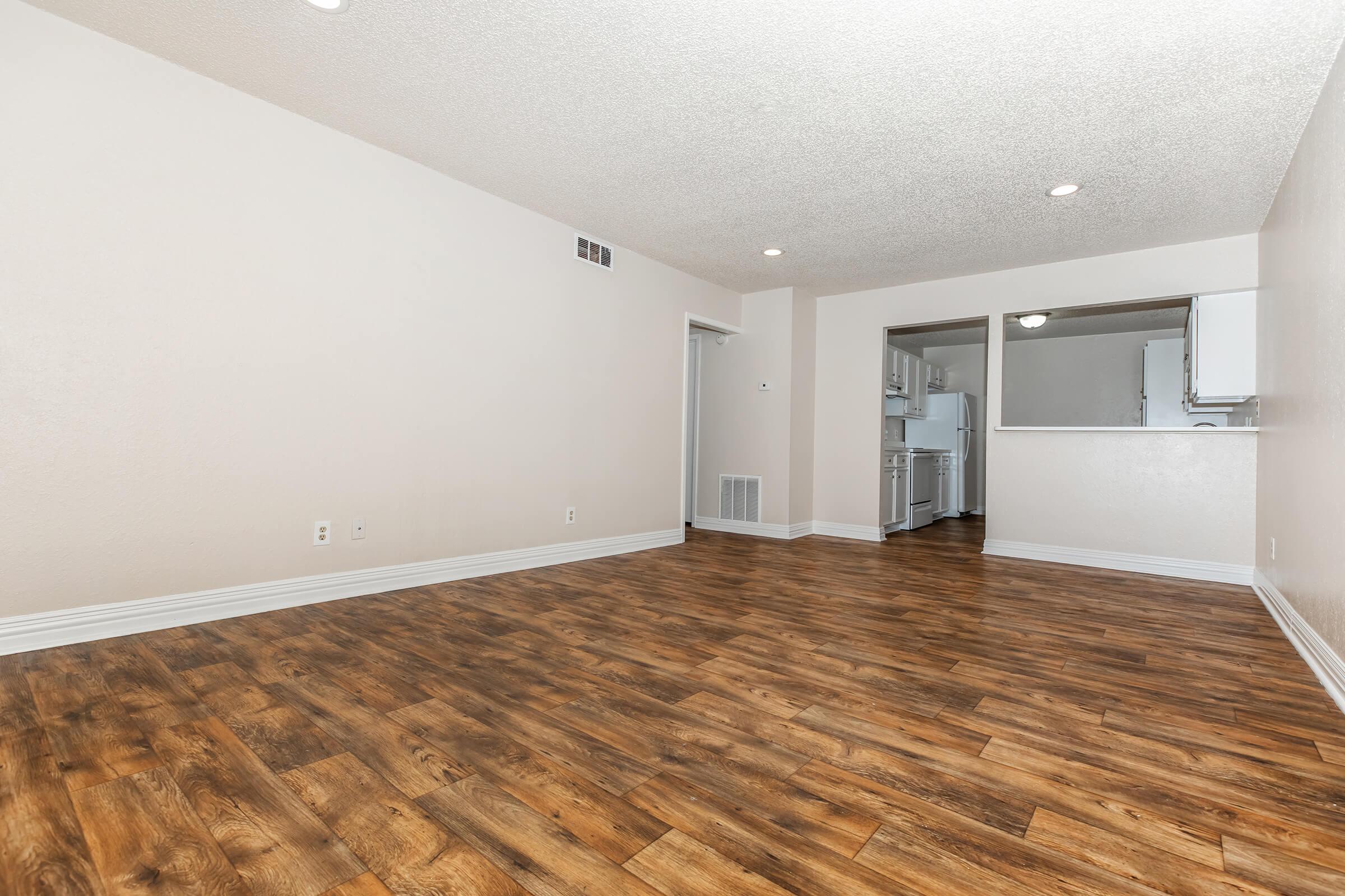 A spacious, empty living area with light-colored walls and a textured ceiling. The floor features rich, dark wood laminate. On the left, a doorway leads to another room, while an open kitchen is visible in the background through a pass-through. The overall ambiance is bright and inviting.