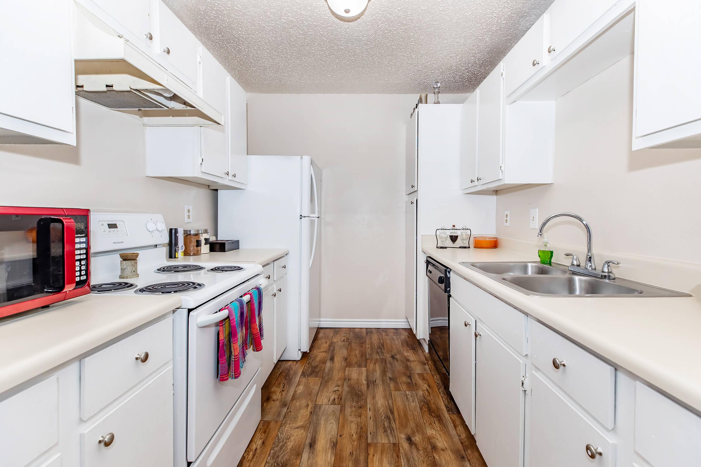 A modern kitchen featuring white cabinetry, a stove with stovetop, a microwave, a refrigerator, and a dual sink. The countertops are clean and organized, with a few kitchen utensils and a colorful towel hanging from the oven. The flooring is a warm wood grain, creating a cozy atmosphere.