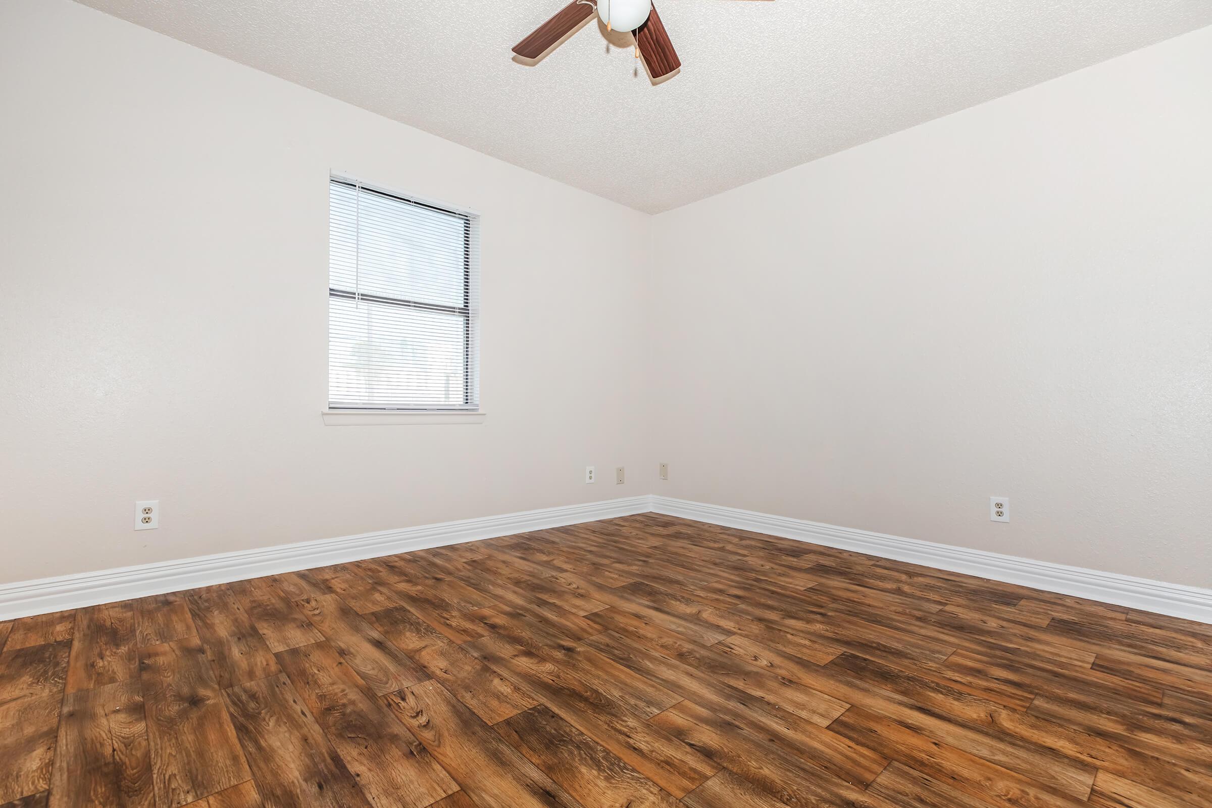 A spacious, empty room with light-colored walls and a wooden floor. There is a single window with horizontal blinds on the far wall, and a ceiling fan is visible. The room has a bright and airy feel, suitable for various purposes such as a bedroom or office.