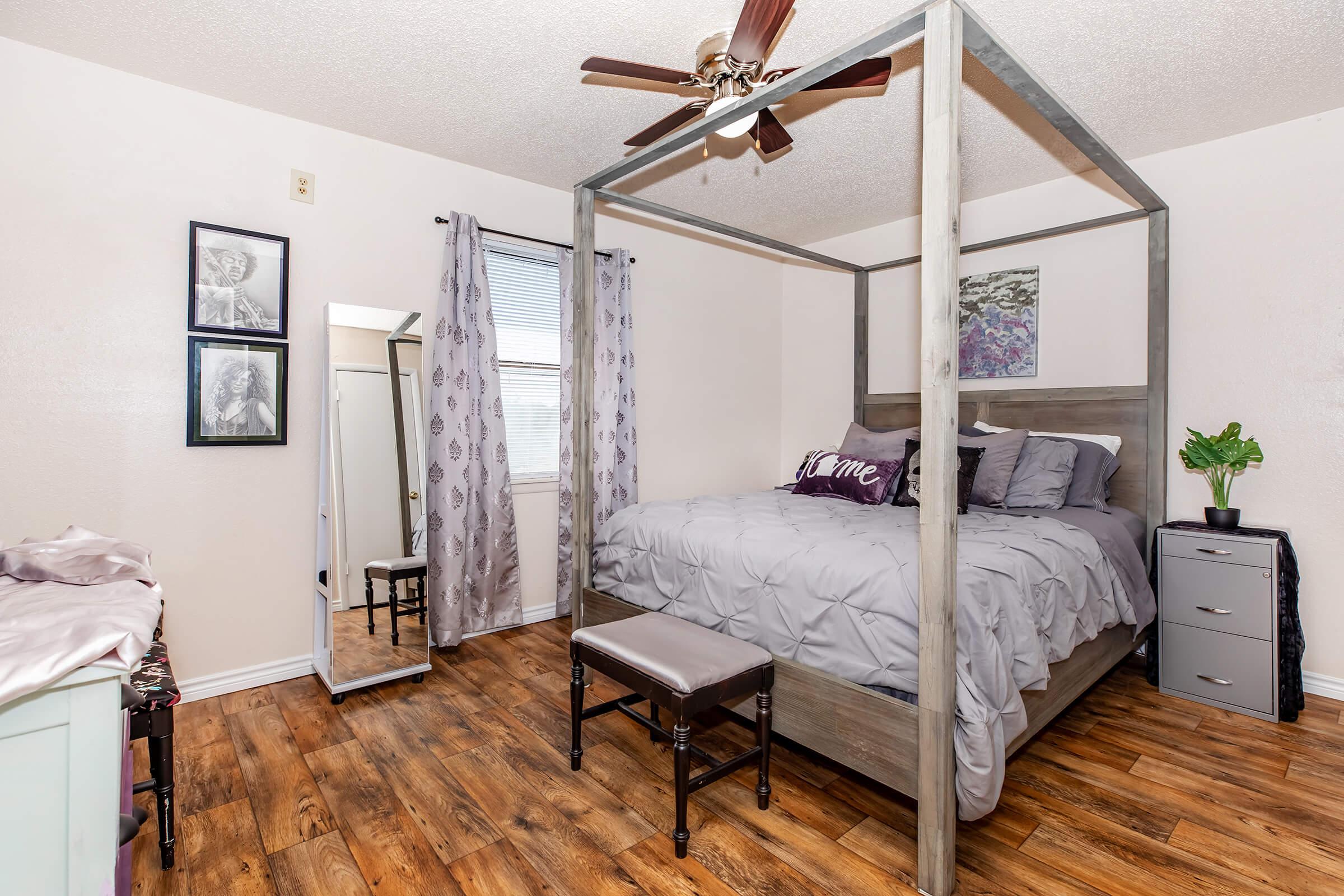 A cozy bedroom featuring a four-poster bed with gray bedding and decorative pillows, a wooden chair, a large mirror, and a small nightstand. Natural light comes through a window with light curtains, and there is a potted plant in the corner. The floor has wood-like flooring, contributing to a warm atmosphere.