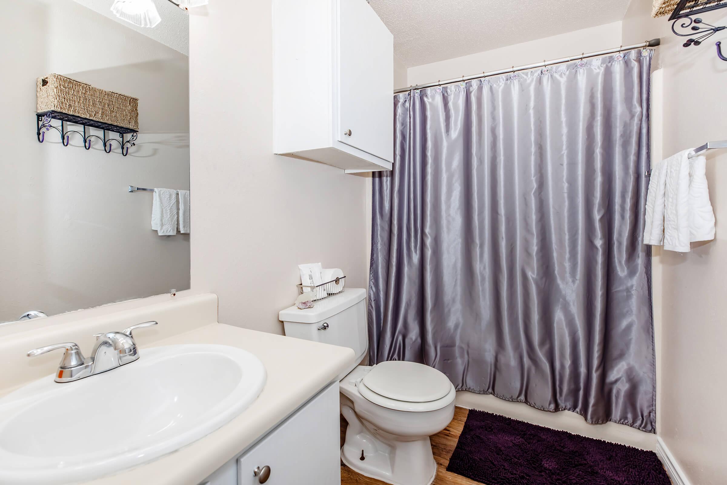 A clean bathroom featuring a white sink and a toilet. The space has light-colored walls, a gray shower curtain, and wooden flooring. There are neatly arranged towels and toiletries on the counter, along with a woven basket on a shelf. A plush purple bath mat complements the decor.