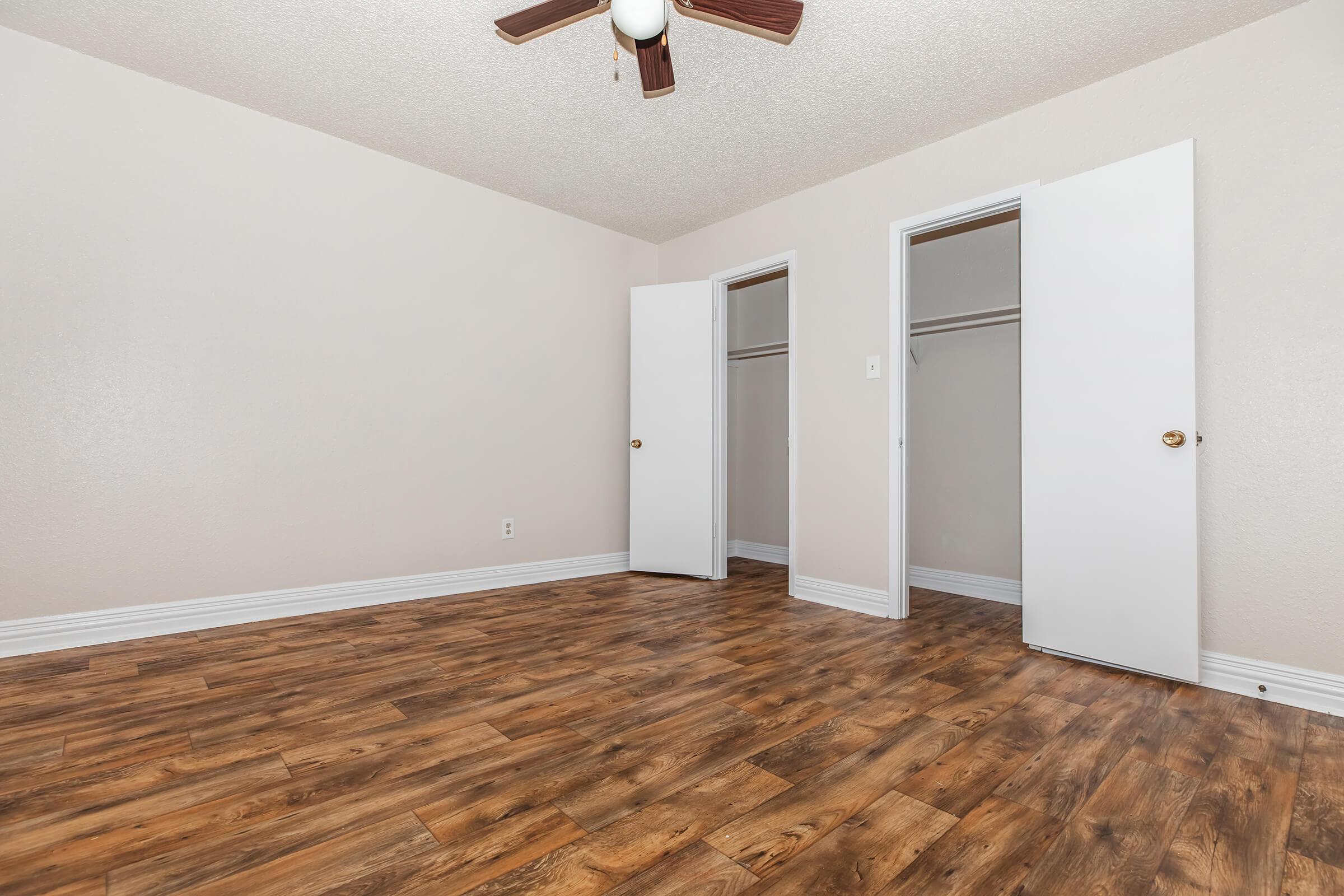 Spacious room with light-colored walls and a ceiling fan. The floor features wood-like laminate, and there are two open closet doors on the right. The room has a clean, neutral aesthetic, ideal for various furnishings and decor. Bright and airy atmosphere with plenty of natural light.