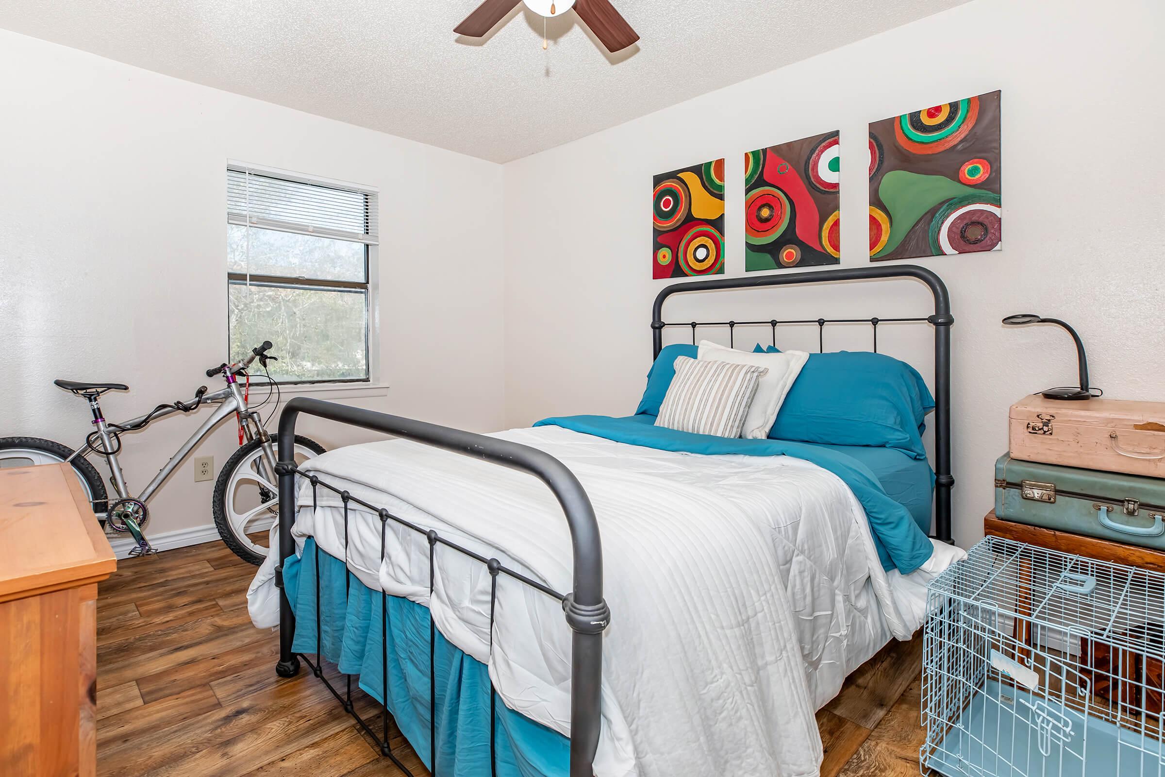 A cozy bedroom featuring a metal-framed bed with blue bedding and decorative pillows. A colorful triptych artwork hangs above the bed. A dresser is visible, along with a bicycle and a pet crate in the corner. Natural light enters through a window, enhancing the inviting atmosphere.