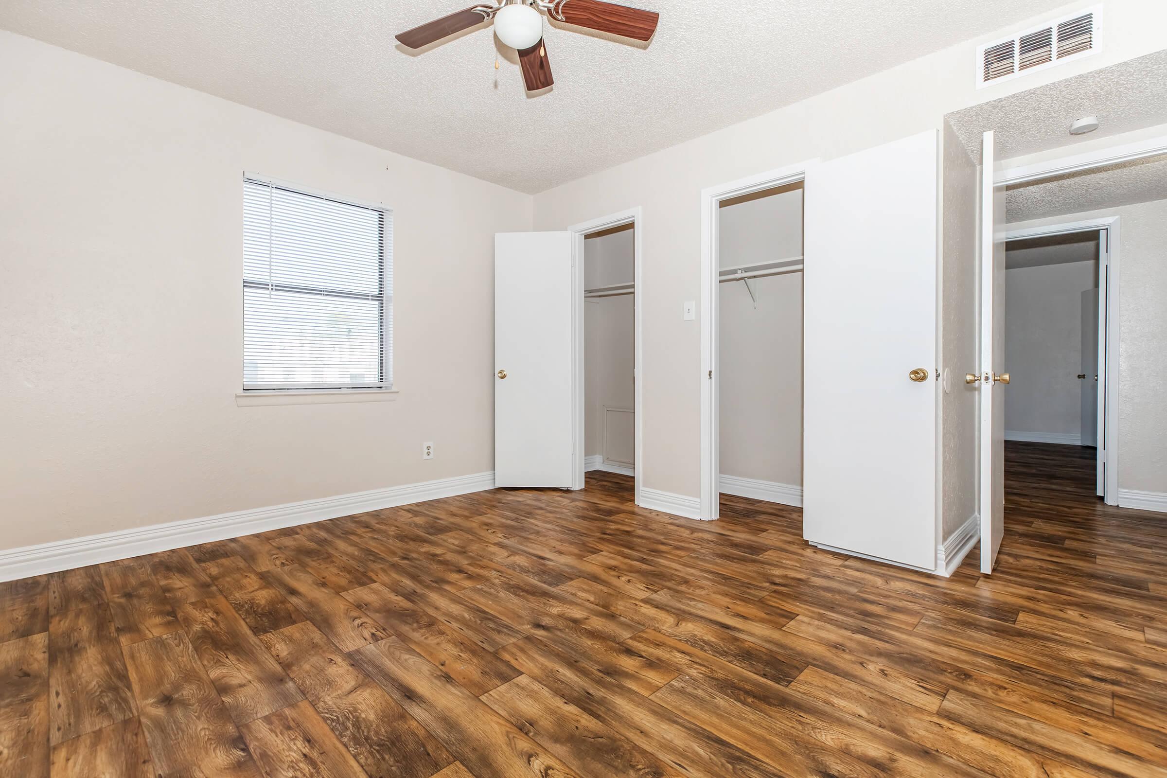 A bright, empty room featuring a ceiling fan, light-colored walls, and large windows. There are two white closet doors open on the left, revealing the closets inside. The floor has a wood-like finish, adding warmth to the space. The room is well-lit, showing a clean and spacious layout.