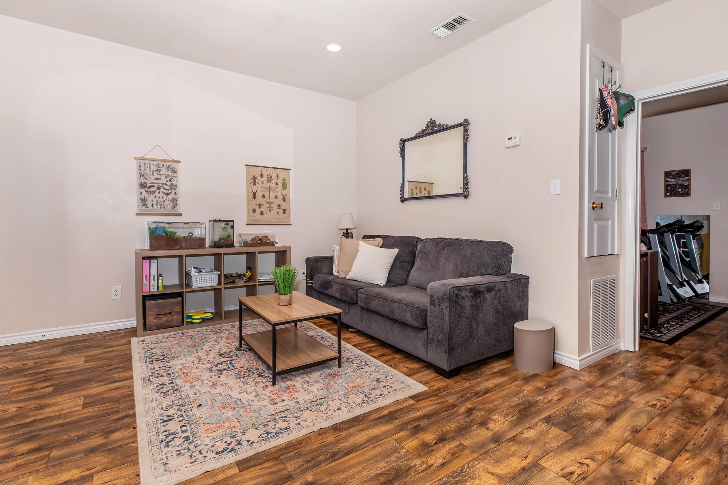 A cozy living room featuring a gray couch with white pillows, a wooden coffee table, and a decorative rug. On the wall hang two framed botanical prints, and a mirror reflects the space. In the corner, there's a small plant and a shelf with books. A door leads to another room with exercise equipment visible.