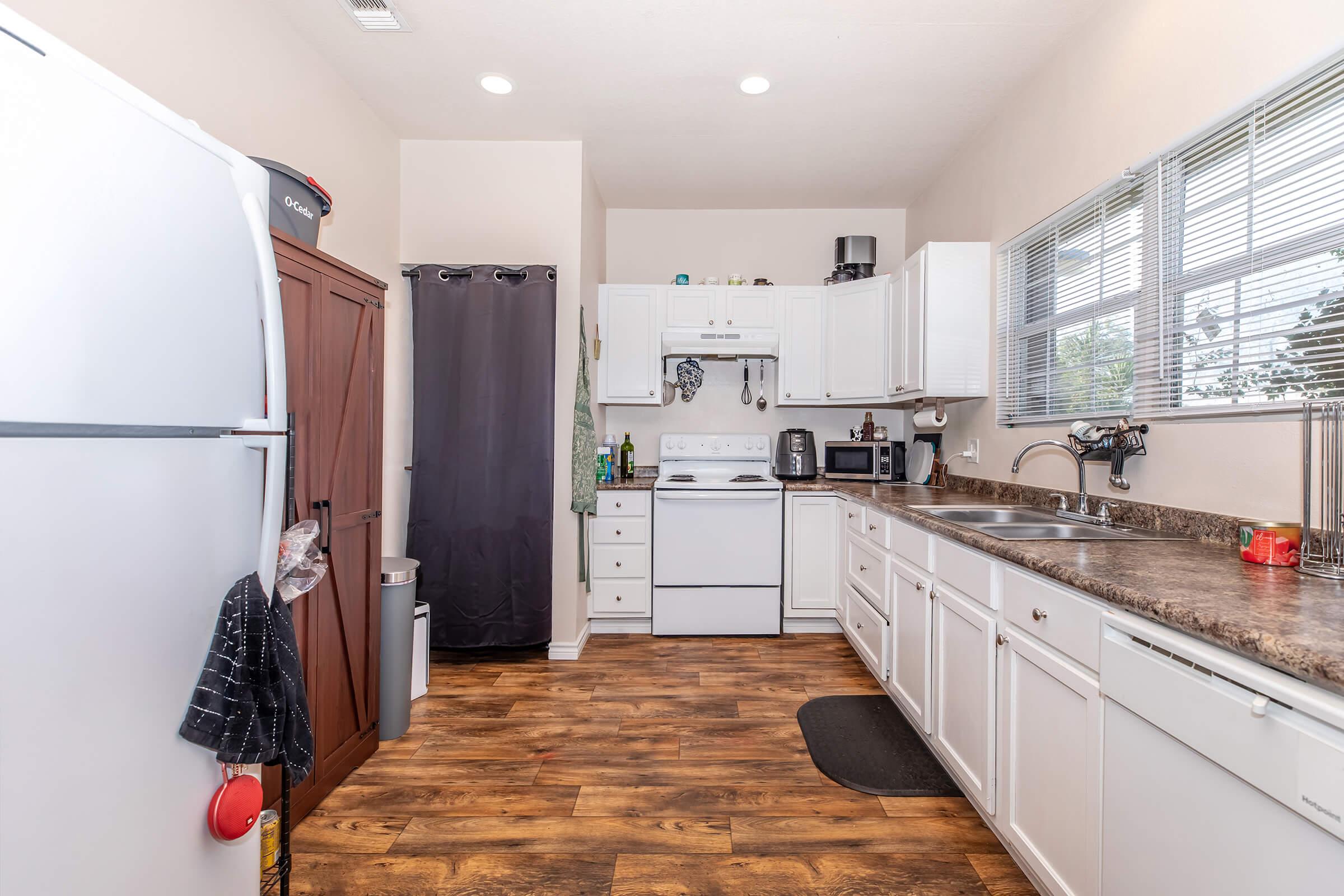 A bright, modern kitchen featuring a refrigerator, stove, sink, and countertops. White cabinets provide storage, and a window allows natural light to enter. The floor has a warm wooden appearance, and small appliances are visible on the counter. A dark curtain partially covers a doorway.