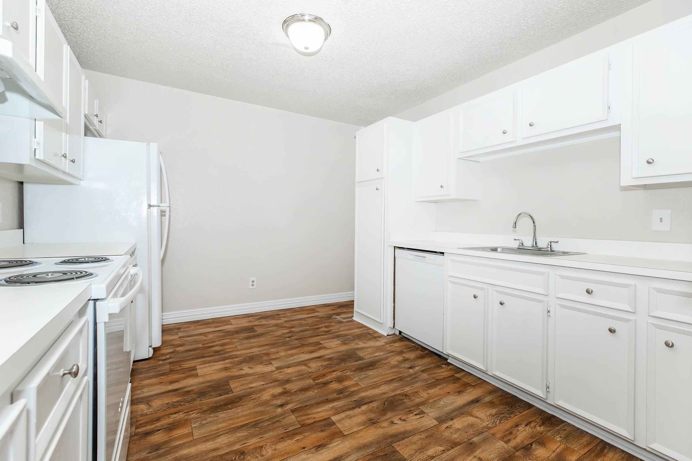 A clean and modern kitchen featuring white cabinetry, a sink, and appliances including a refrigerator and stove. The flooring is a warm wood-like laminate, and the walls are light-colored, creating a bright and open atmosphere. Simplistic design with ample counter space.