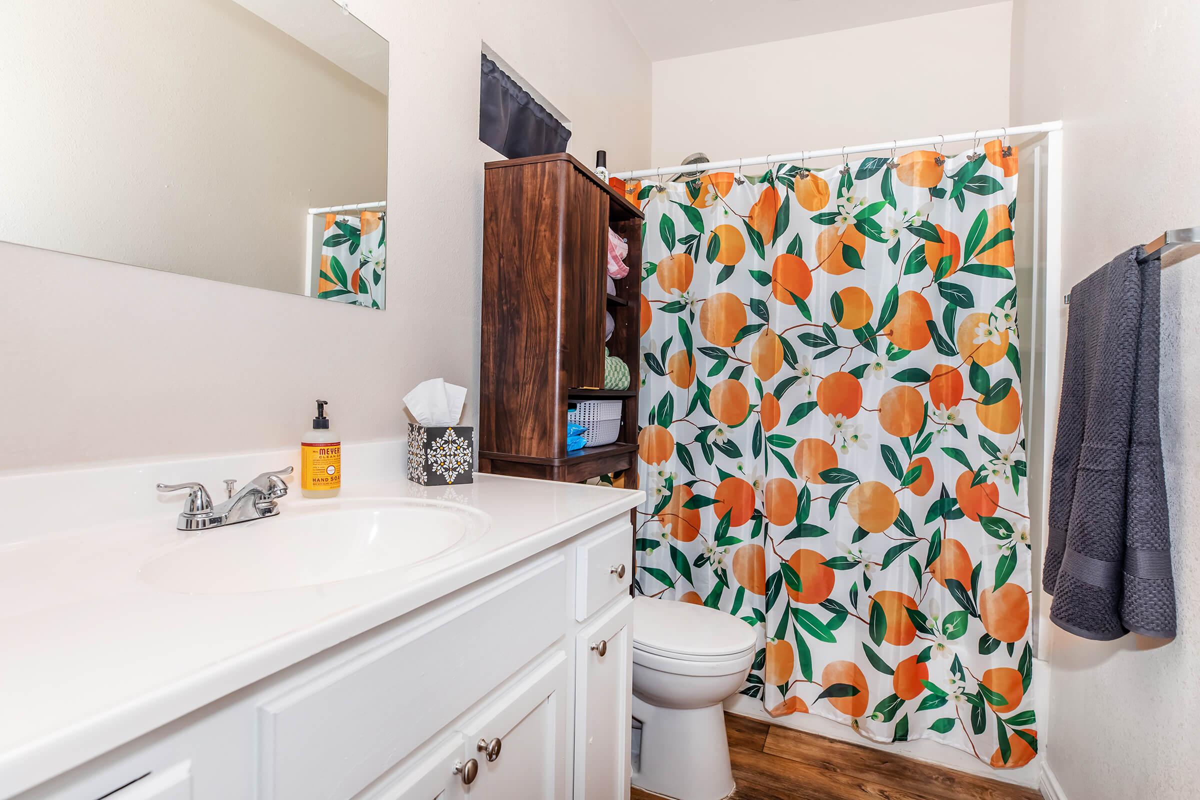 A bright bathroom featuring a white vanity with a sink, a large mirror, and a toilet. The space is decorated with an orange-themed shower curtain adorned with leaves and bright oranges. There is a wooden shelf holding toiletries and a rolled towel on a wall hook, complementing the vibrant decor.