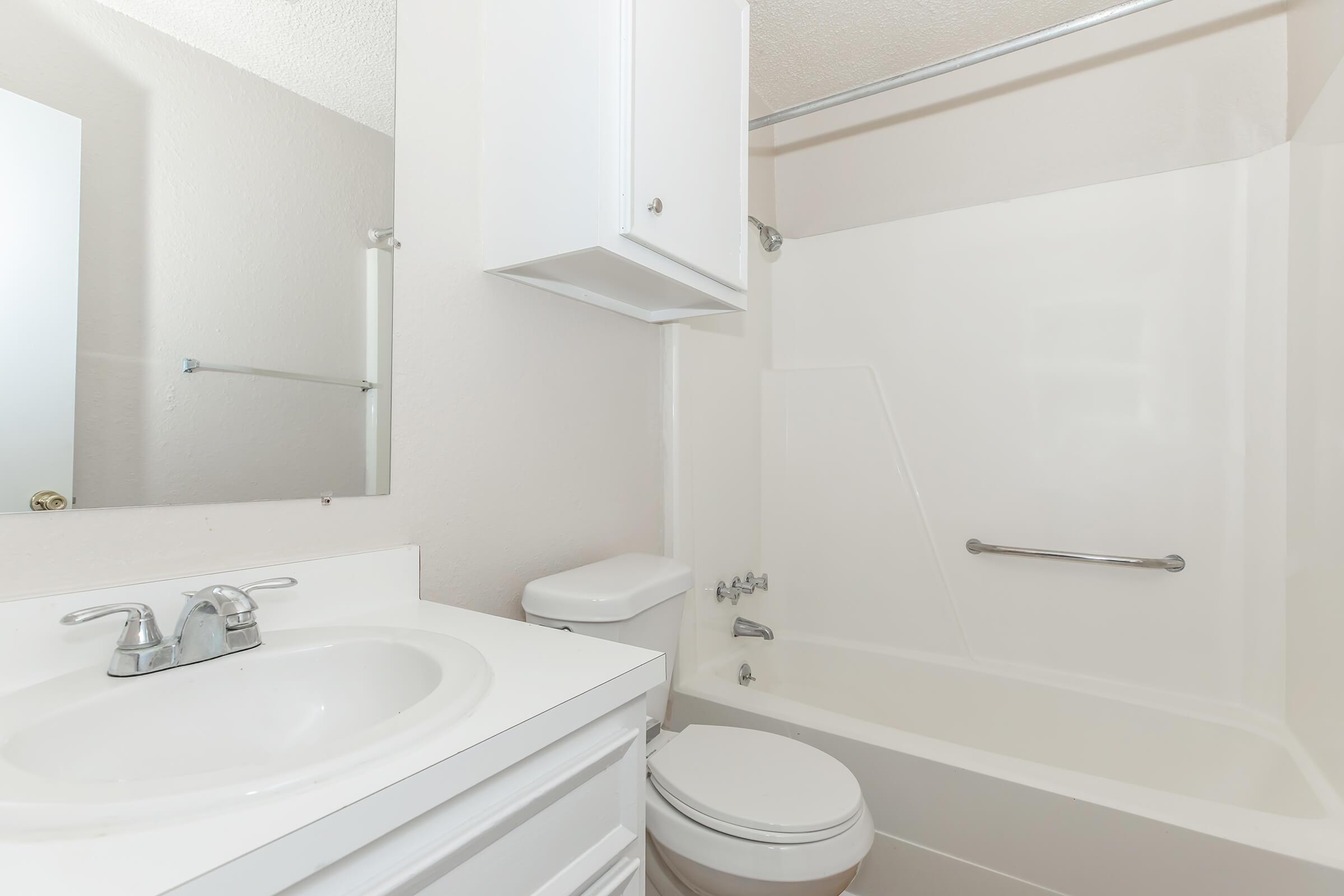 A clean and modern bathroom featuring a white bathtub with a shower, a white toilet, and a sink with a chrome faucet. The walls are light-colored, and there's a small cabinet above the sink. A mirror is mounted on the wall, reflecting a spacious, well-lit environment.