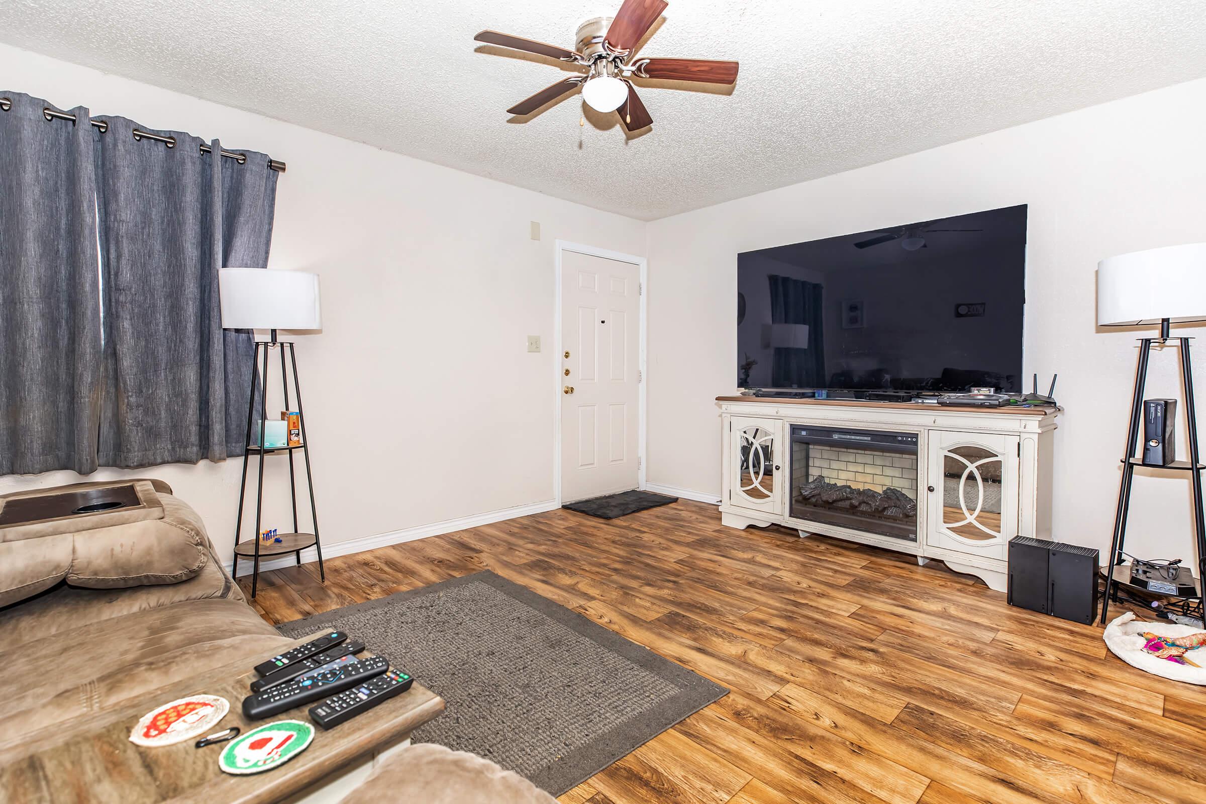 Cozy living room with a ceiling fan, a large flat-screen TV mounted on a light-colored entertainment center, and a comfortable couch. There are two stylish floor lamps, gray curtains on the window, and a front door visible in the background. The flooring is wood-like, adding warmth to the space.