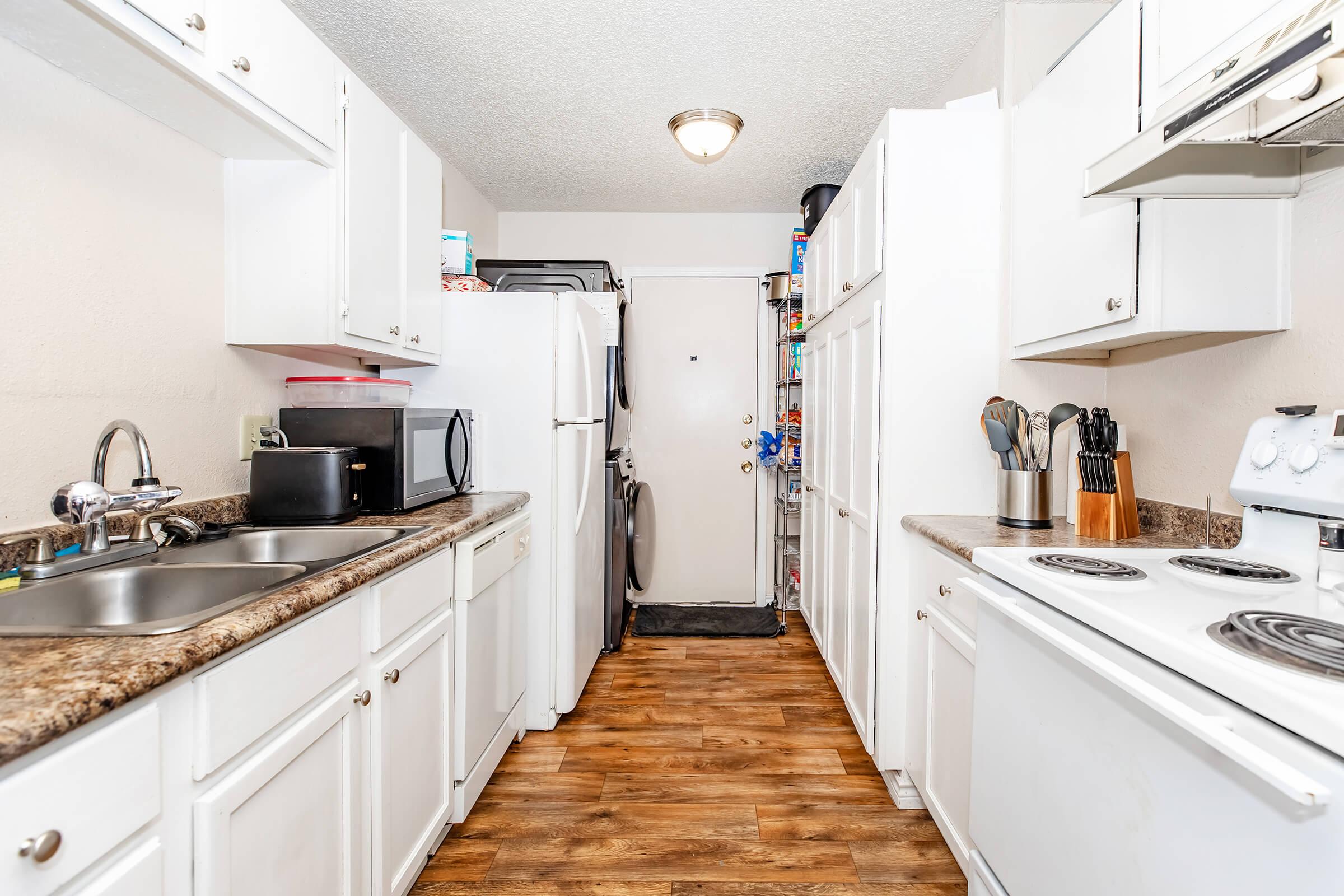 A narrow kitchen featuring white cabinets and a dark countertop. It includes a double sink, a microwave, a refrigerator, and a stove. The walls are painted light, and the floor has a wood-like finish. The door at the end leads to an exit, with a few appliances and kitchen tools visible on the countertops.