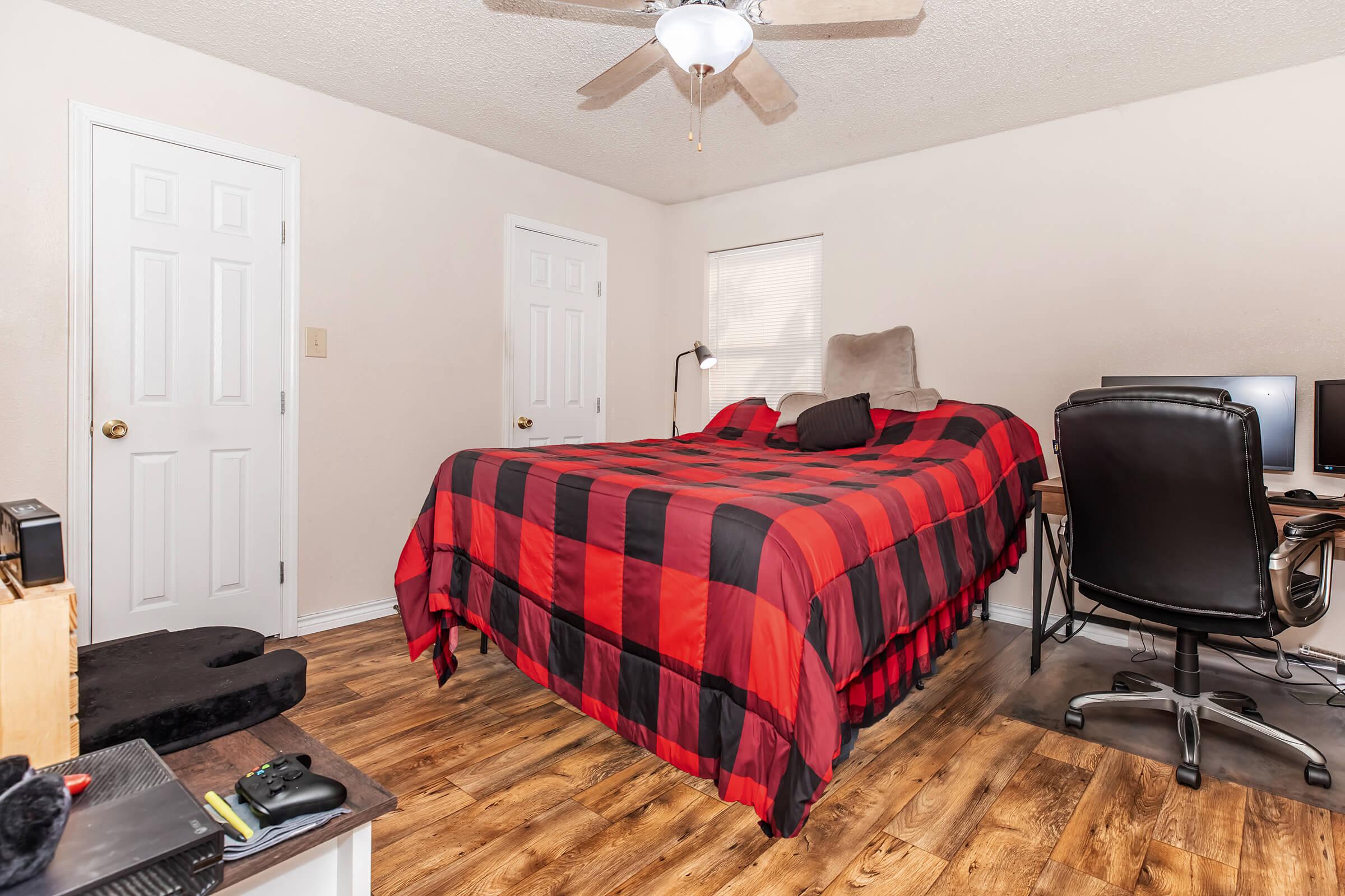 A cozy bedroom featuring a large bed with a red and black checkered comforter. There are two white doors, a window with light filtering through, and a small workspace with two computer monitors and a black chair. The floor is wood laminate, adding warmth to the space.