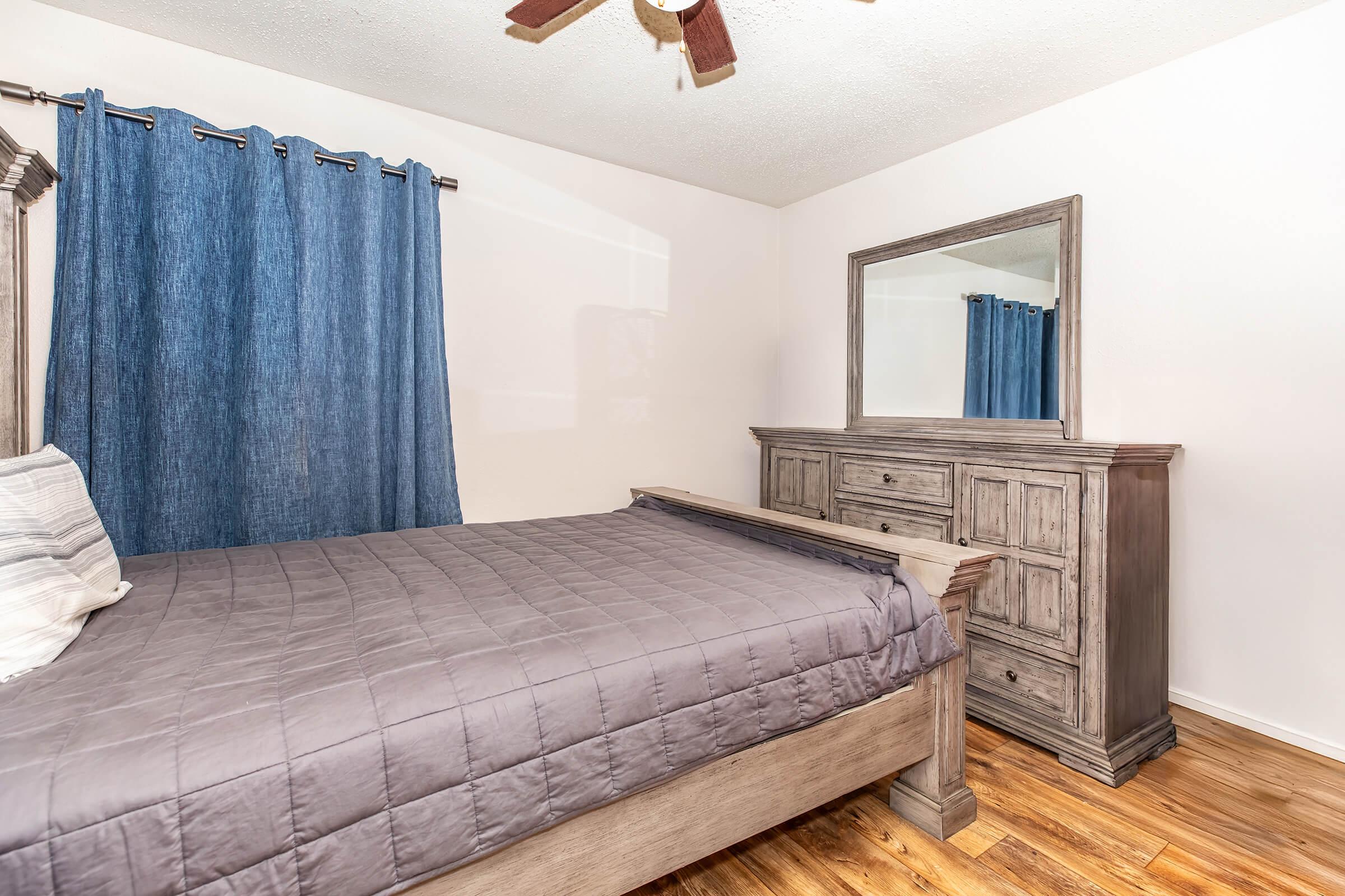 A cozy bedroom featuring a wooden bed frame with a gray quilt, a large dresser with a mirror, and blue curtains hanging over a window. The floor is made of wood, creating a warm, inviting atmosphere.
