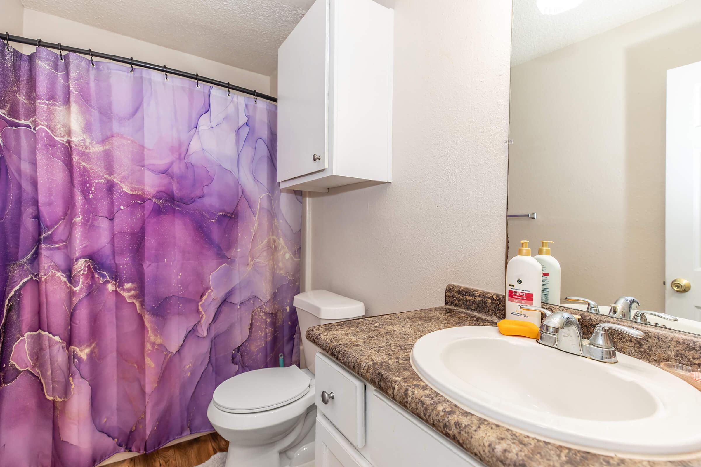 A modern bathroom featuring a purple and gold abstract shower curtain, a white vanity with a sink, and toiletries on the counter. The decor is simple, with neutral walls and natural wood flooring, creating a clean and inviting space.