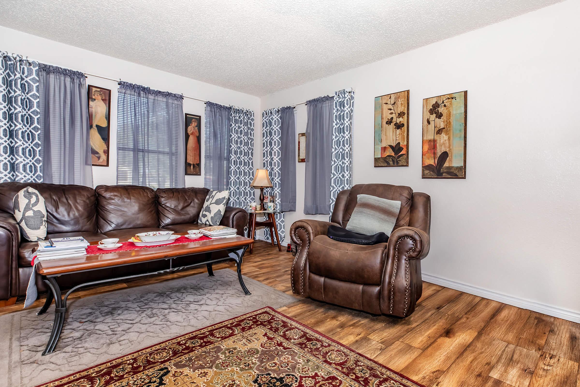 A cozy living room featuring a brown leather sofa, an armchair, a coffee table with decor items, and a patterned area rug. Windows are dressed with gray curtains, and the walls display framed artwork, adding a touch of elegance to the warm, inviting atmosphere.