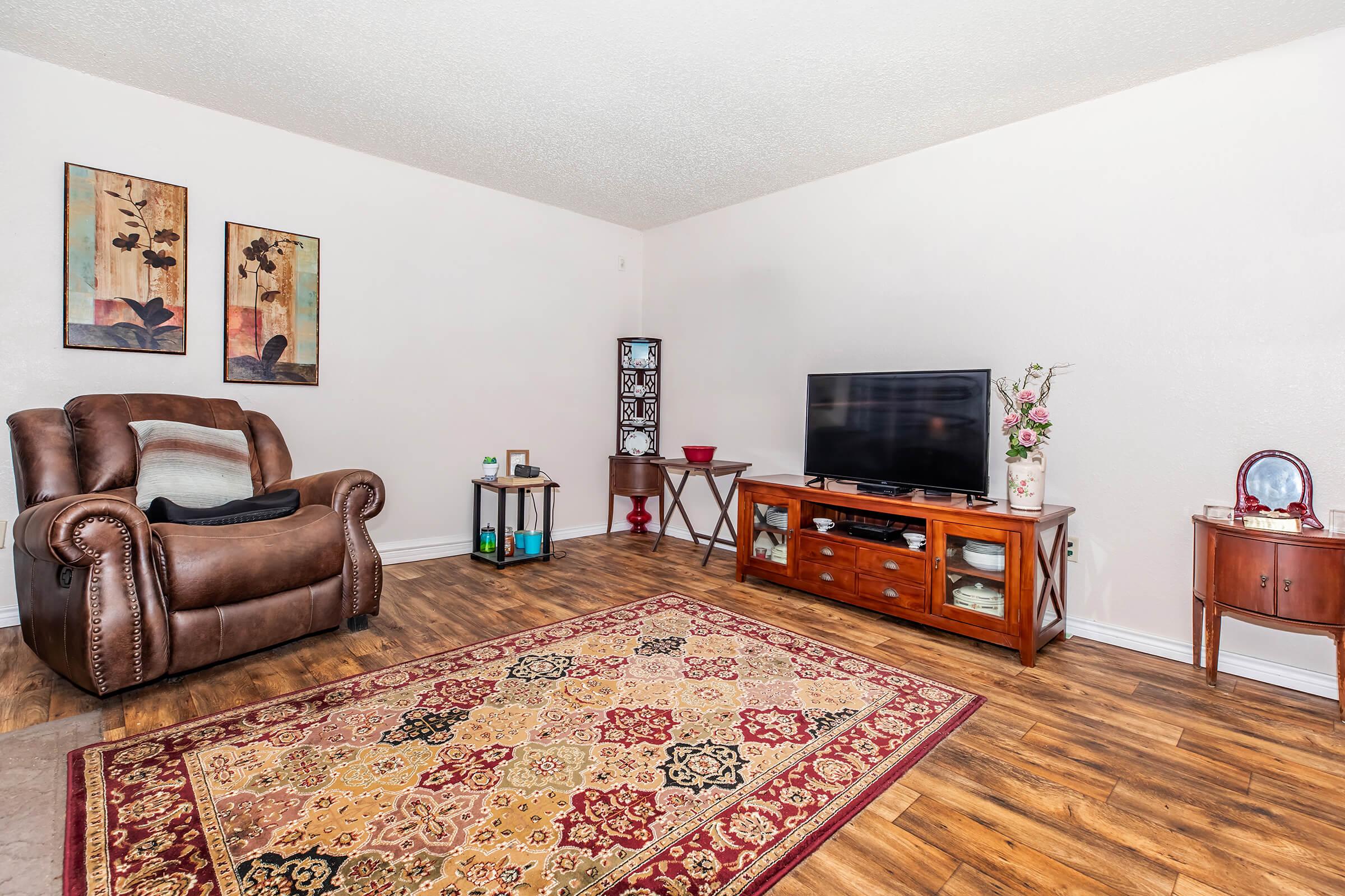 A cozy living room featuring a brown recliner chair, a wooden TV stand with a flat-screen television, and a decorative side table. The floor is covered with a patterned rug, and wall art with floral themes adds warmth to the space. Natural light enhances the inviting atmosphere.