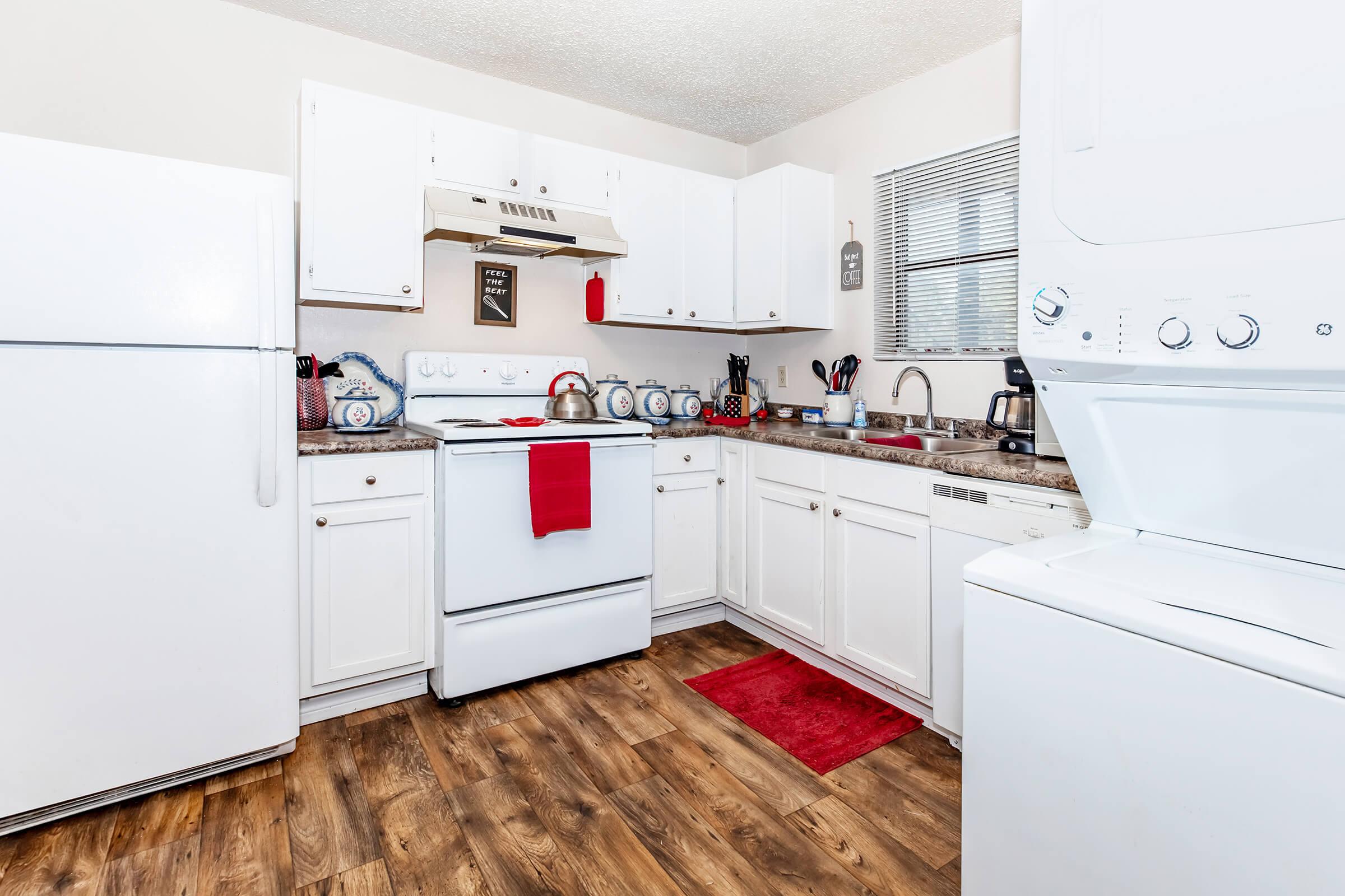 A bright, modern kitchen featuring white cabinetry, a white refrigerator, and a stovetop with a range hood. There are various kitchen utensils and blue dishes on the countertop, along with a red dish towel hanging from the oven. A washing machine and dryer are visible on the right side, and wood-like flooring completes the look.