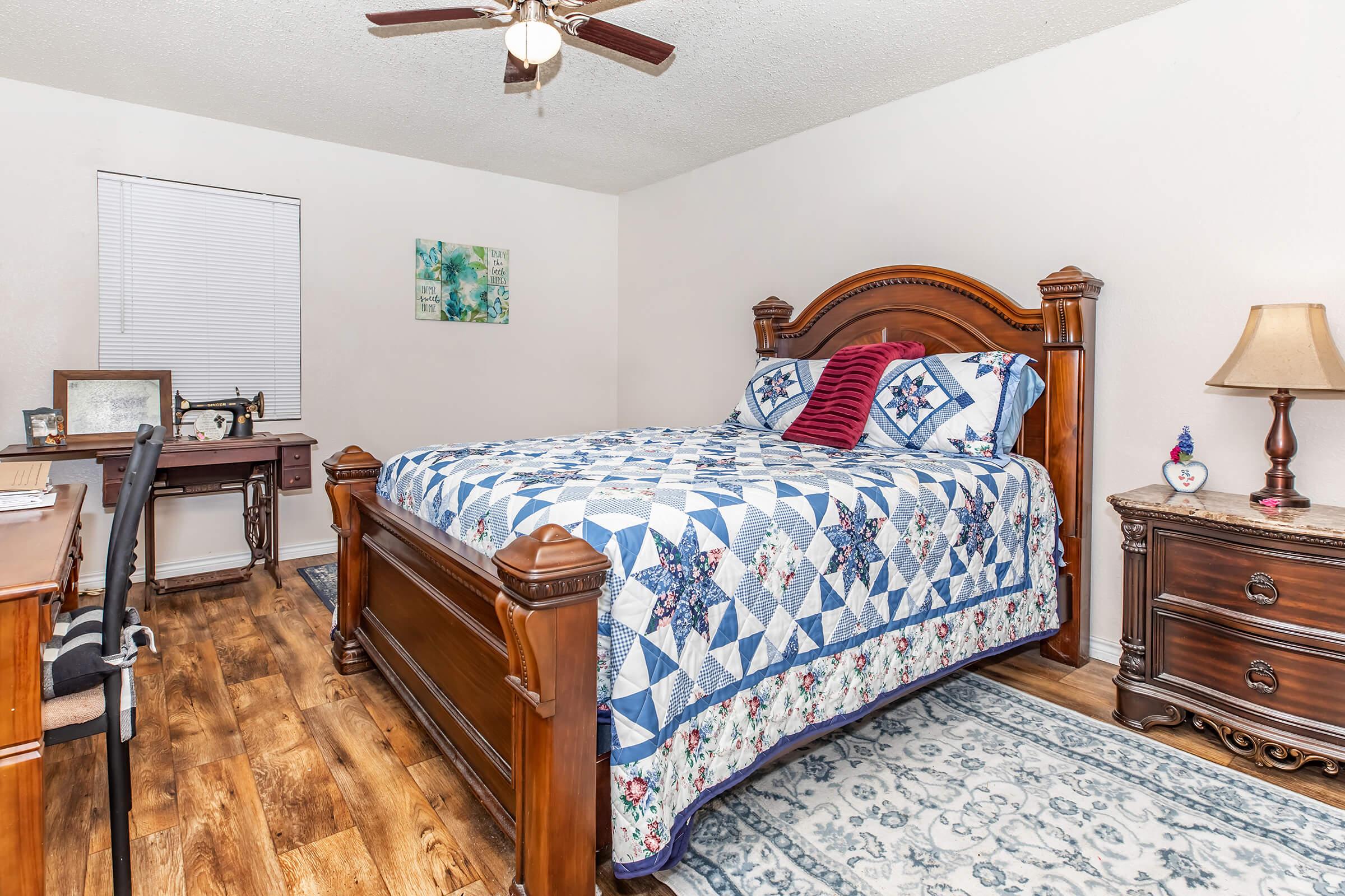 A cozy bedroom featuring a wooden bed with a blue and white quilt. There’s a desk and chair to the left, a lamp on a nightstand, and a decorative rug on the floor. The walls are painted light, with a window providing natural light and a piece of art hanging decoratively.