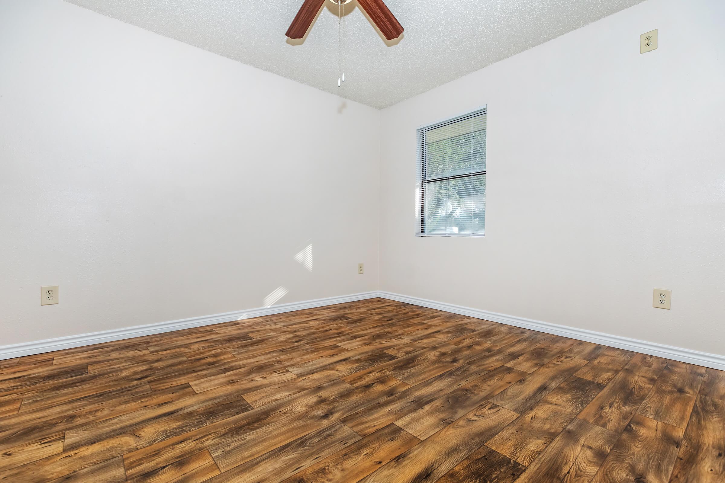 Empty room with light-colored walls and a wooden floor. A ceiling fan with wooden blades hangs from the ceiling, and there's a window letting in natural light. The space is mostly bare, suggesting it is ready for furniture or decoration.