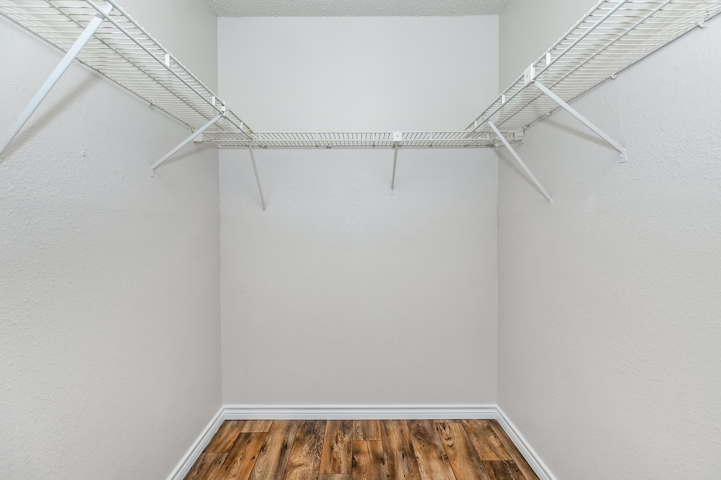 Empty closet with white wire shelving on two walls, light gray painted walls, and a wooden floor. The space is clean and organized, ideal for storage or organizing items.