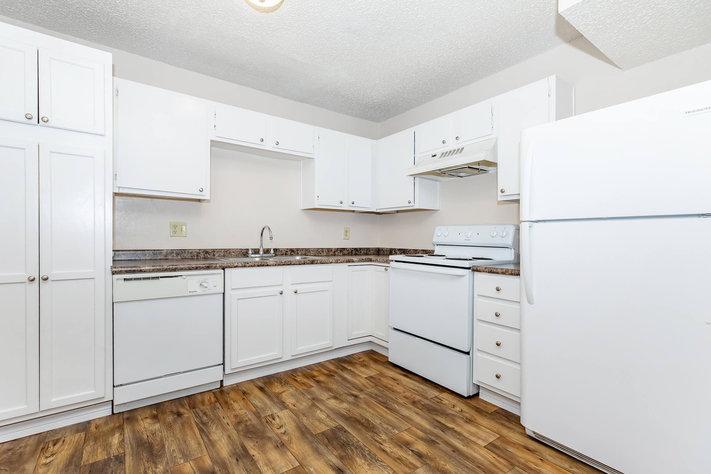 A clean, modern kitchen featuring white cabinets, a white refrigerator, and a white stove. It includes a dishwasher, a sink, and a countertop with dark speckled design. The floor is wood-like laminate, and the walls are painted a neutral color, creating a bright and inviting atmosphere.