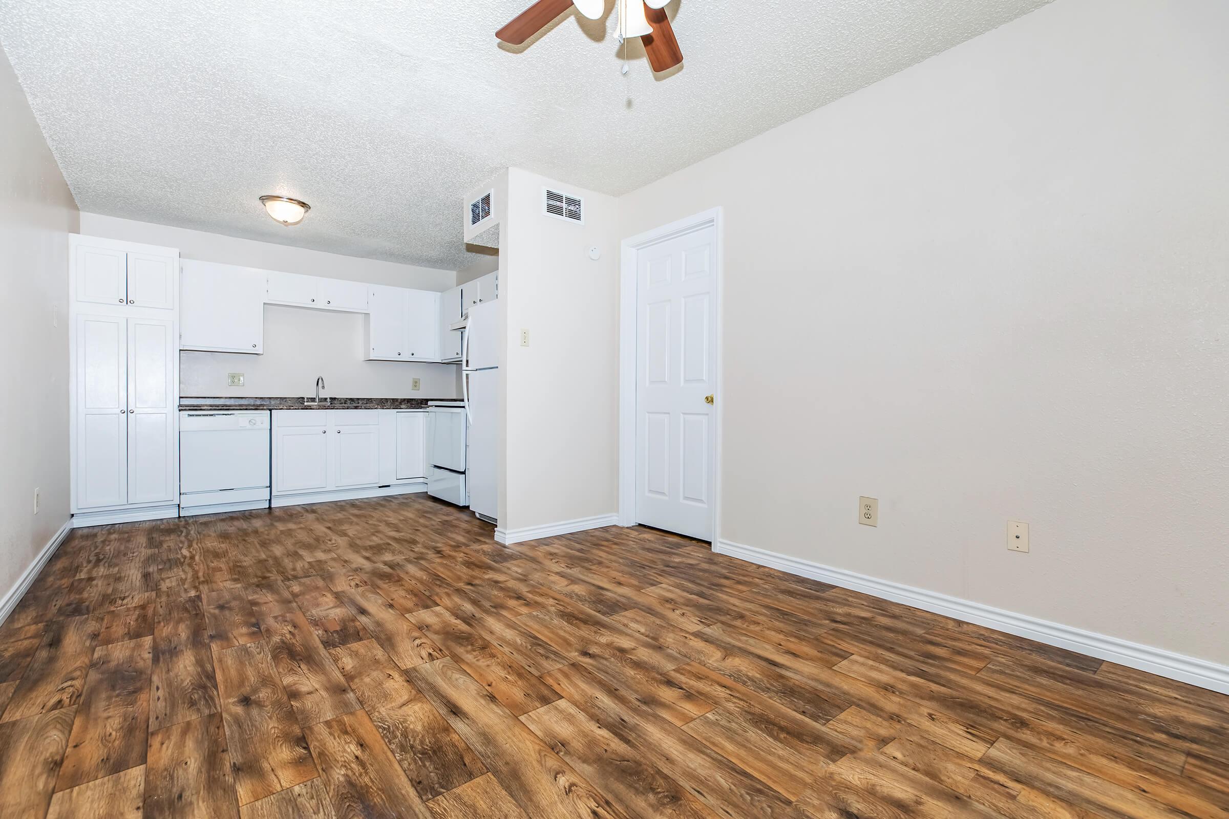A small, clean kitchen area with white cabinetry, a refrigerator, and a stove. The floor is made of light-colored wood laminate. The walls are painted neutral tones, and there's a ceiling fan. A doorway leads to another room, and the space is well-lit with natural light.