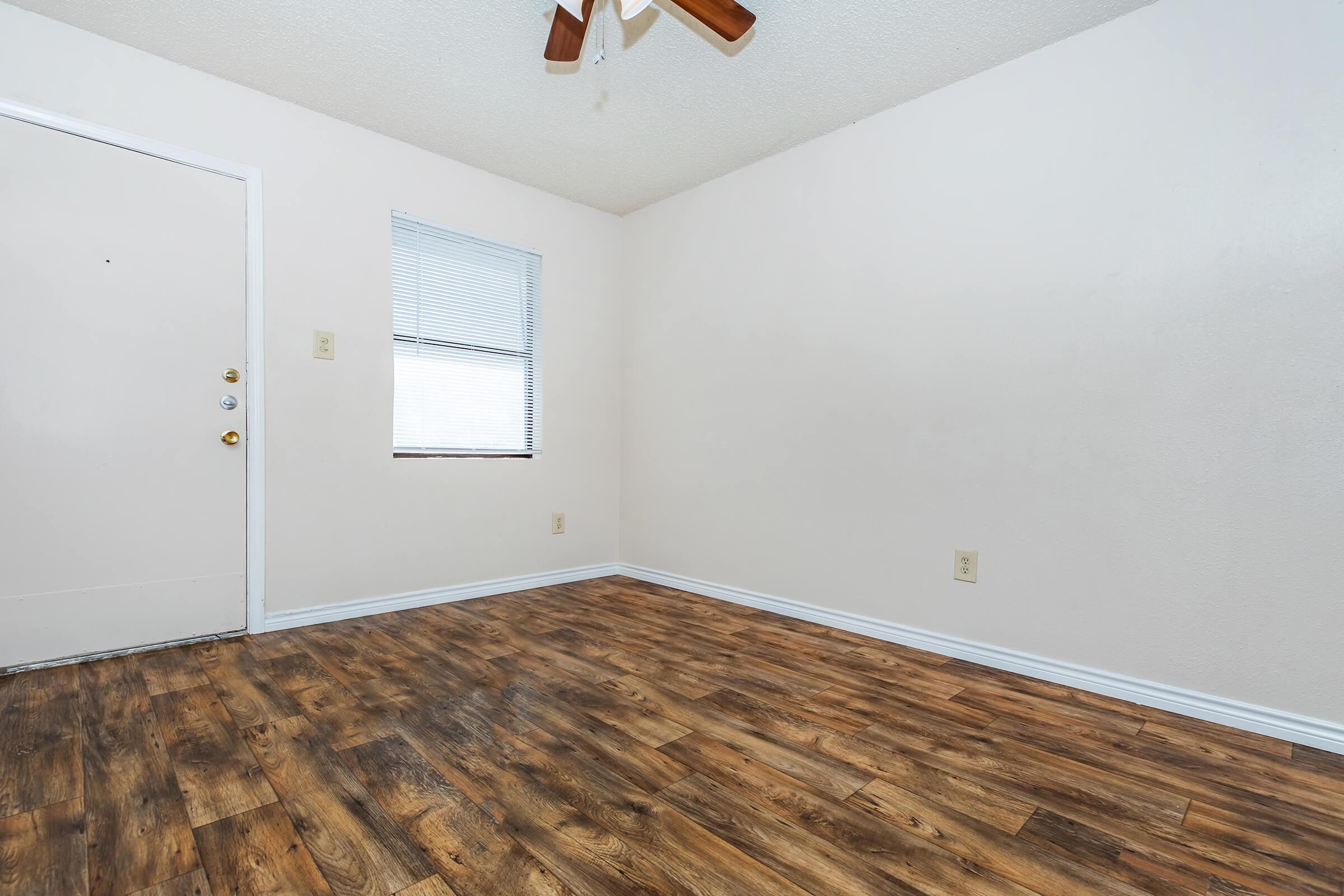 Empty room with light-colored walls and a wooden floor. There is a door on the left side and a small window with blinds on the right. A ceiling fan is visible, adding a casual touch to the space. The room has a bright, airy feel, suitable for various uses like a living area, office, or bedroom.
