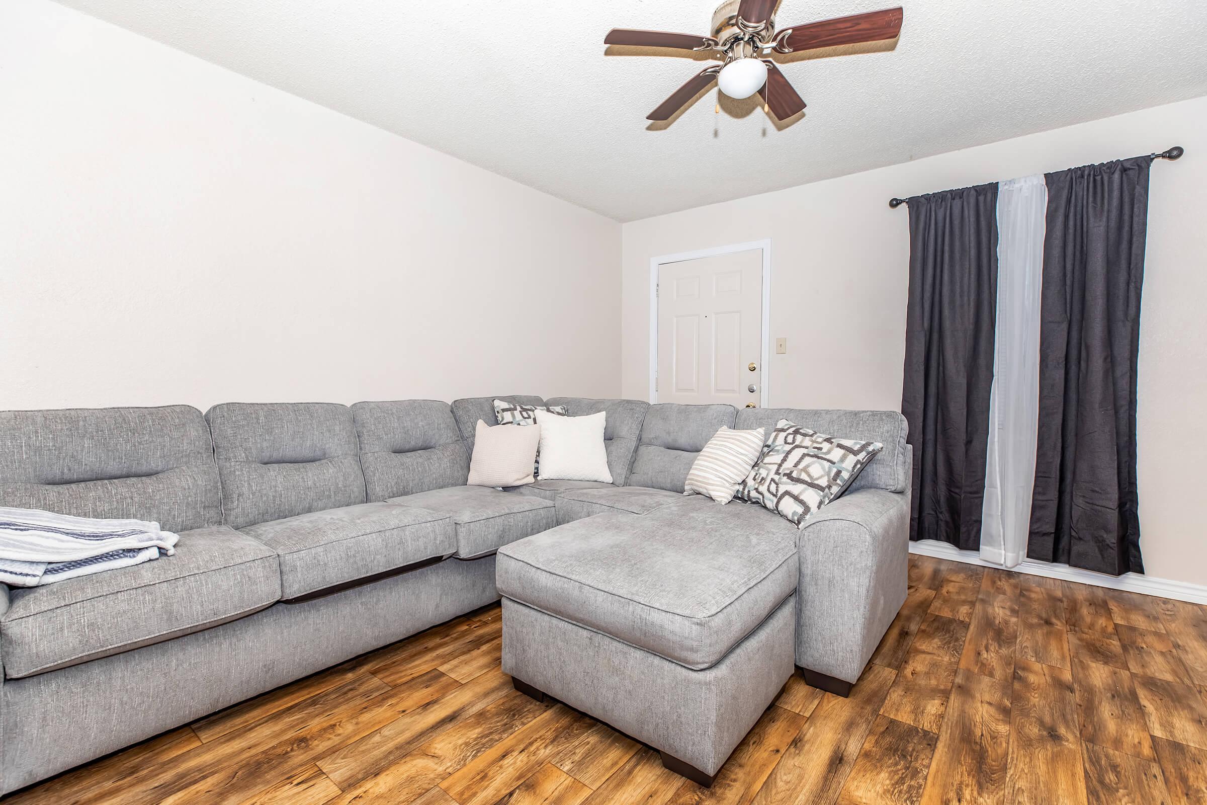 A modern living room featuring a grey sectional sofa with decorative pillows, a carpeted wooden floor, and a ceiling fan. There is a door in the background and a pair of contrasting curtains on the side window. The space has a cozy, inviting atmosphere.