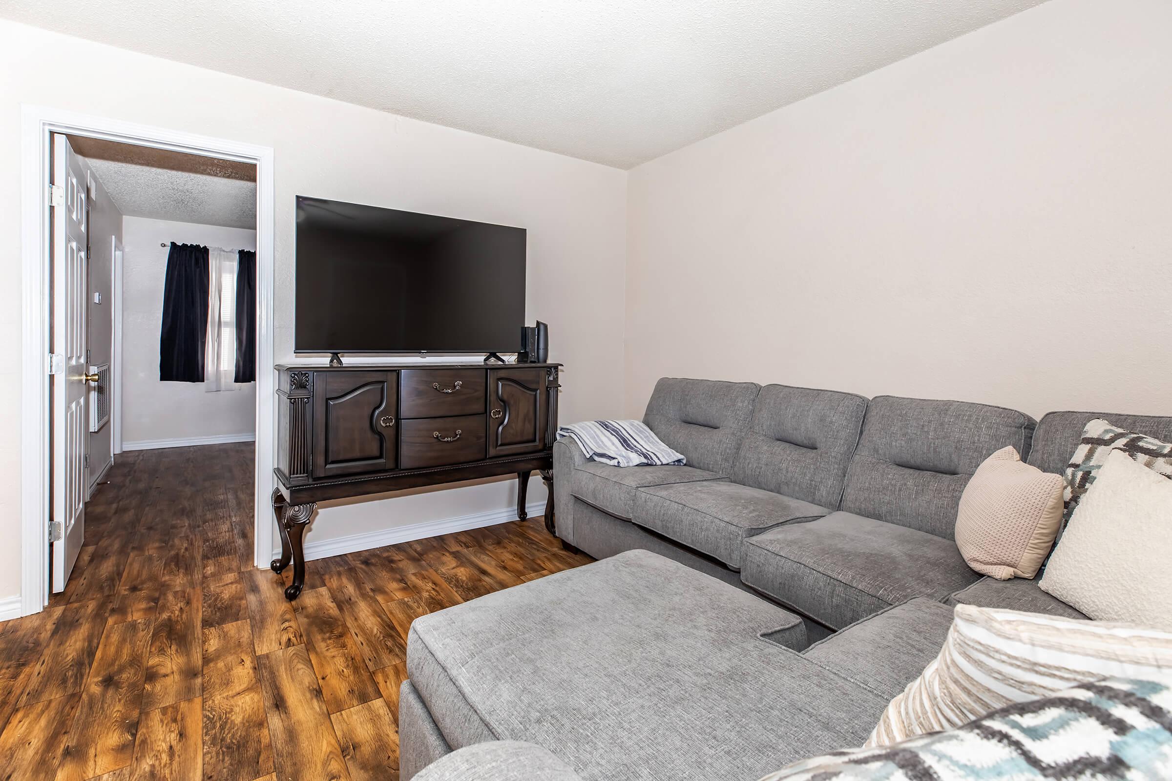 A cozy living room featuring a gray sectional sofa and a large flat-screen TV mounted on a dark wooden cabinet. The floor is made of dark wood, and there is a doorway leading to another room with a window covered by black curtains. Soft pillows and decorative cushions add comfort to the space.