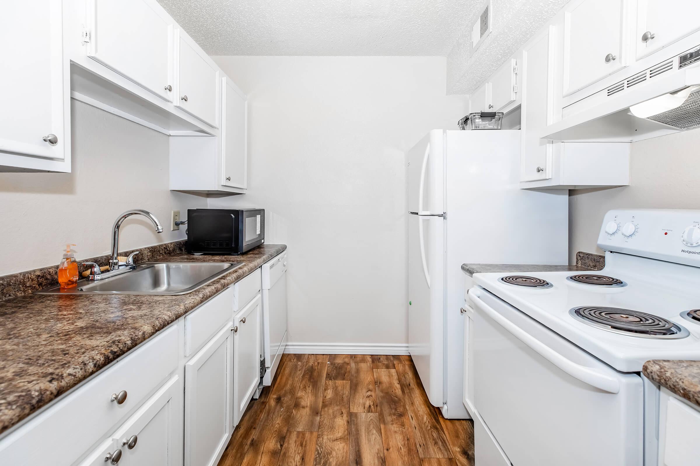 A clean, modern kitchen featuring white cabinets, a dark countertop, a stainless steel sink, a microwave, a white refrigerator, and a white stove with an oven. The floor has a wood-like finish, and the walls are painted in a light color, creating a bright and inviting space.