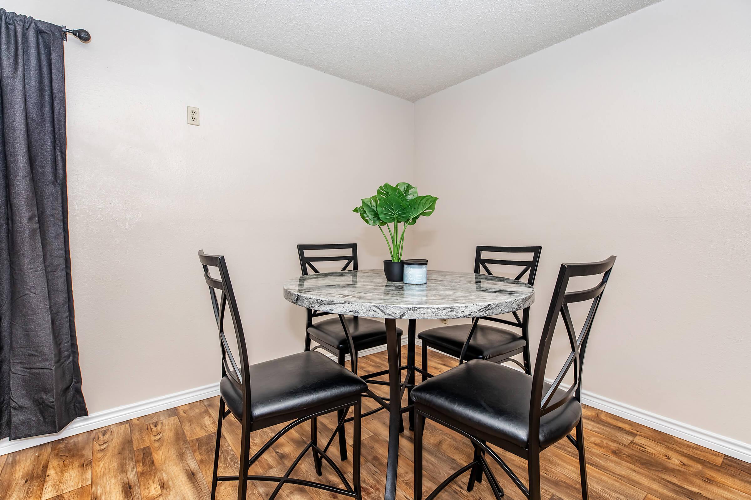 A small dining area featuring a round marble table surrounded by four black chairs. A green potted plant is centered on the table. The walls are light-colored, and there is a dark curtain hanging on one side. The flooring is wooden, creating a cozy atmosphere.