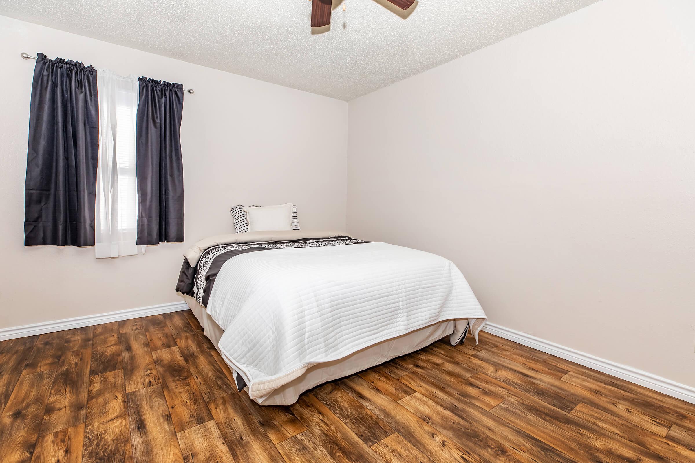 A cozy bedroom featuring a queen-sized bed with white bedding and decorative pillows, set against a light-colored wall. Natural light streams in through a window with sheer and dark curtains. The floor is finished with warm-toned wood, enhancing the inviting atmosphere.