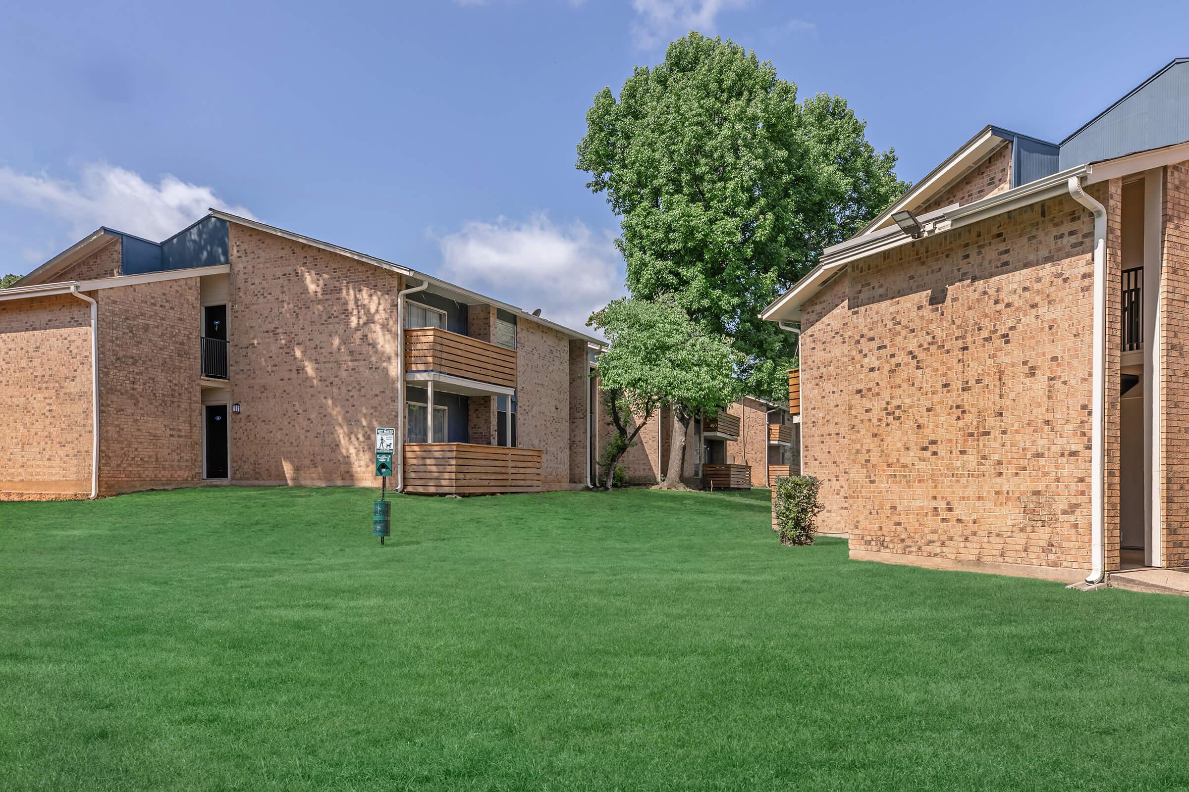 Two residential apartment buildings made of brick, surrounded by well-maintained green lawns. One building features wooden balconies, while the other has a simple façade. A lone tree adds to the scenery under a partly cloudy sky.