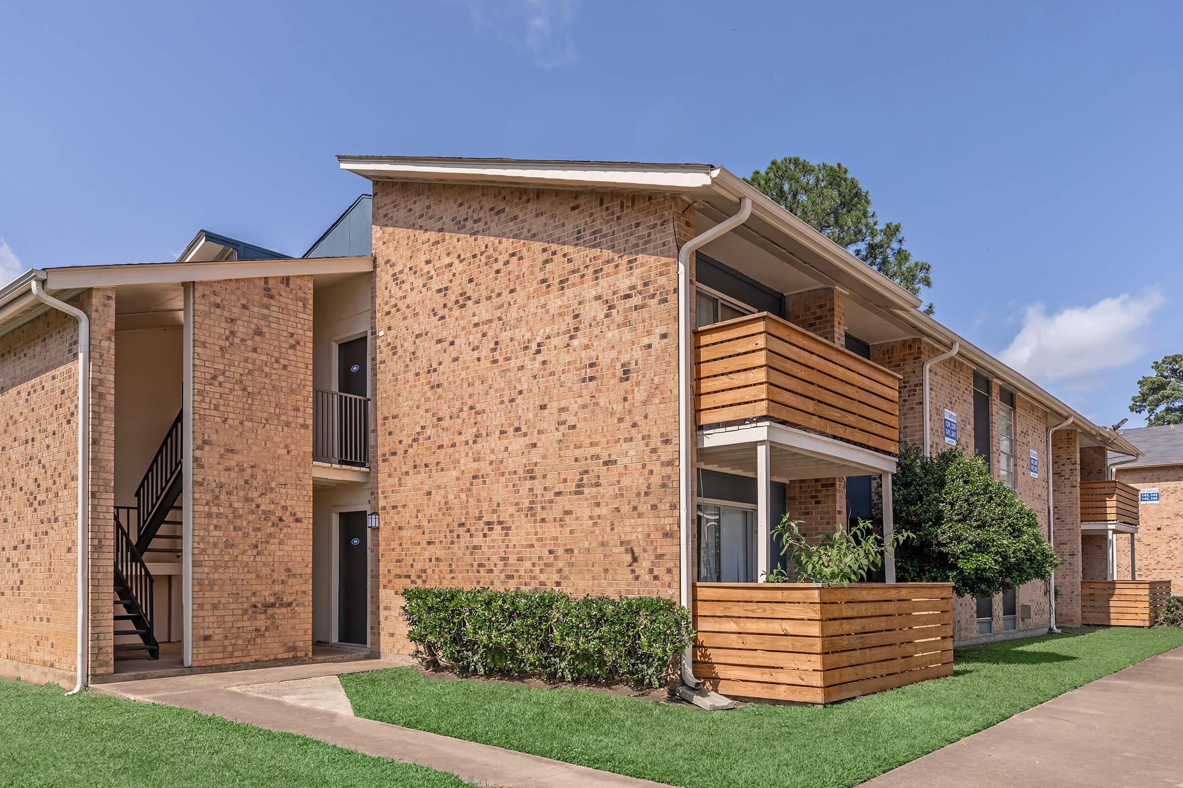 A well-maintained brick apartment complex featuring two stories, balconies with wooden railings, and landscaped grass areas. The entrance includes a staircase, and there are small shrubs along the walkway. The sky is clear with a few clouds.