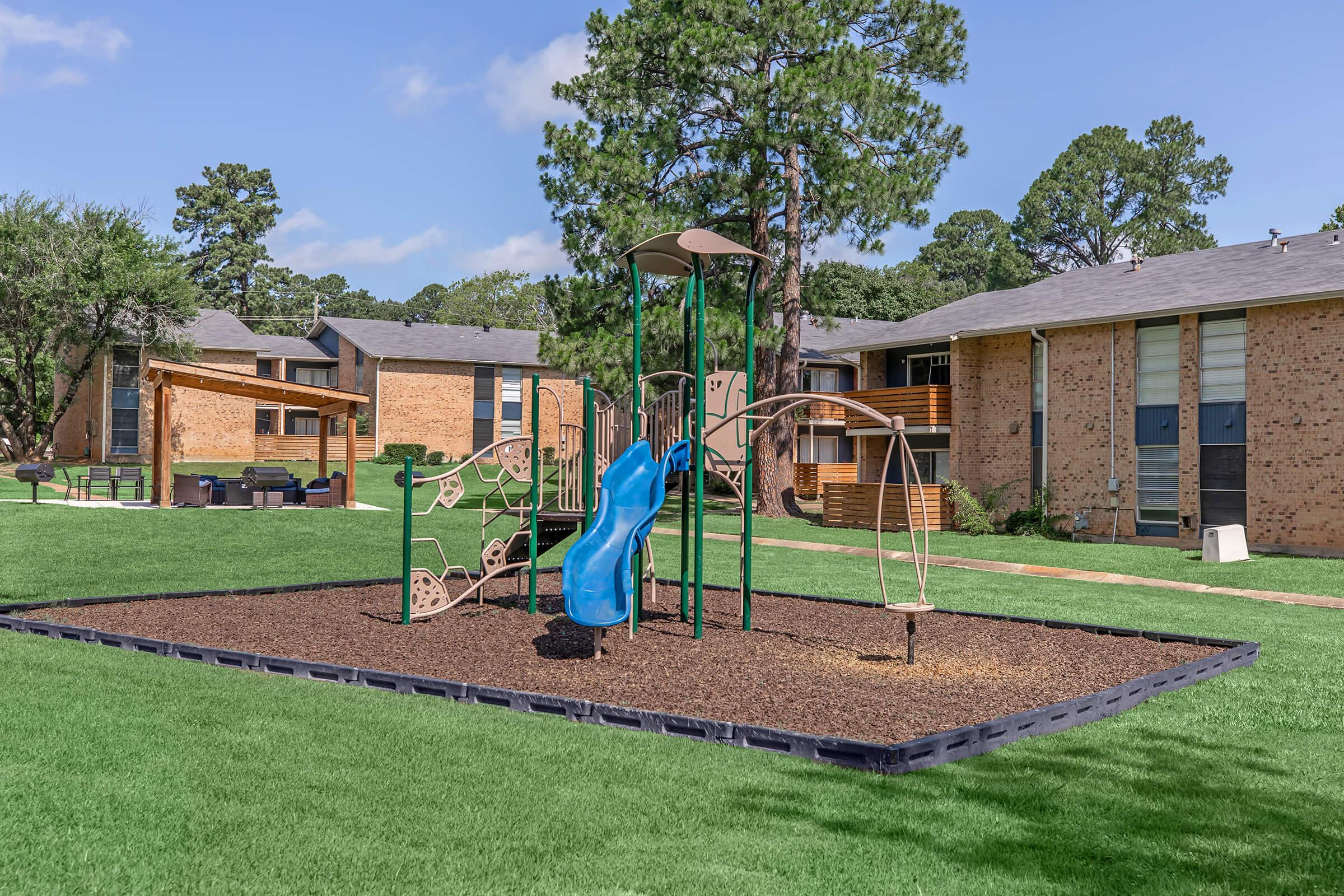 A playground featuring a green and blue slide, climbing structure, and swings, situated on a grassy area surrounded by brick apartment buildings. The sky is clear with a few clouds, and trees provide shade in the background.