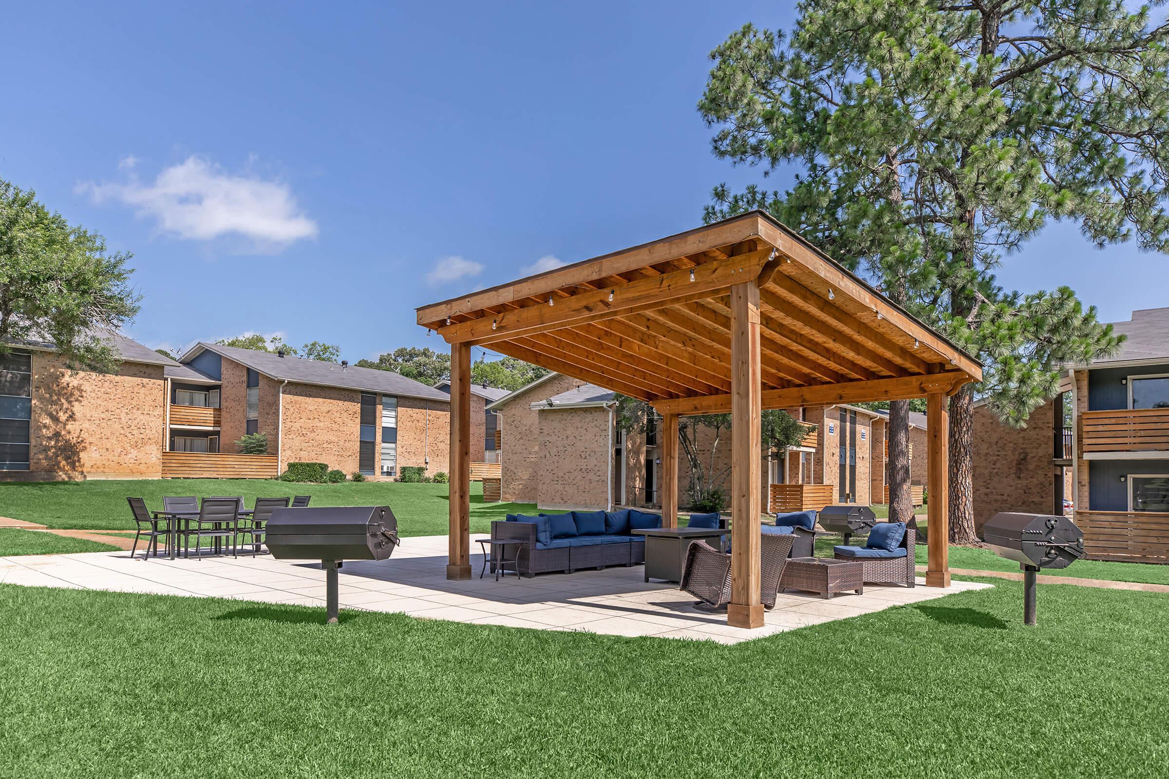 A spacious outdoor patio with a wooden pergola, featuring comfortable seating and a dining table. Two grills are positioned nearby on a well-maintained lawn, surrounded by residential buildings under a clear blue sky.