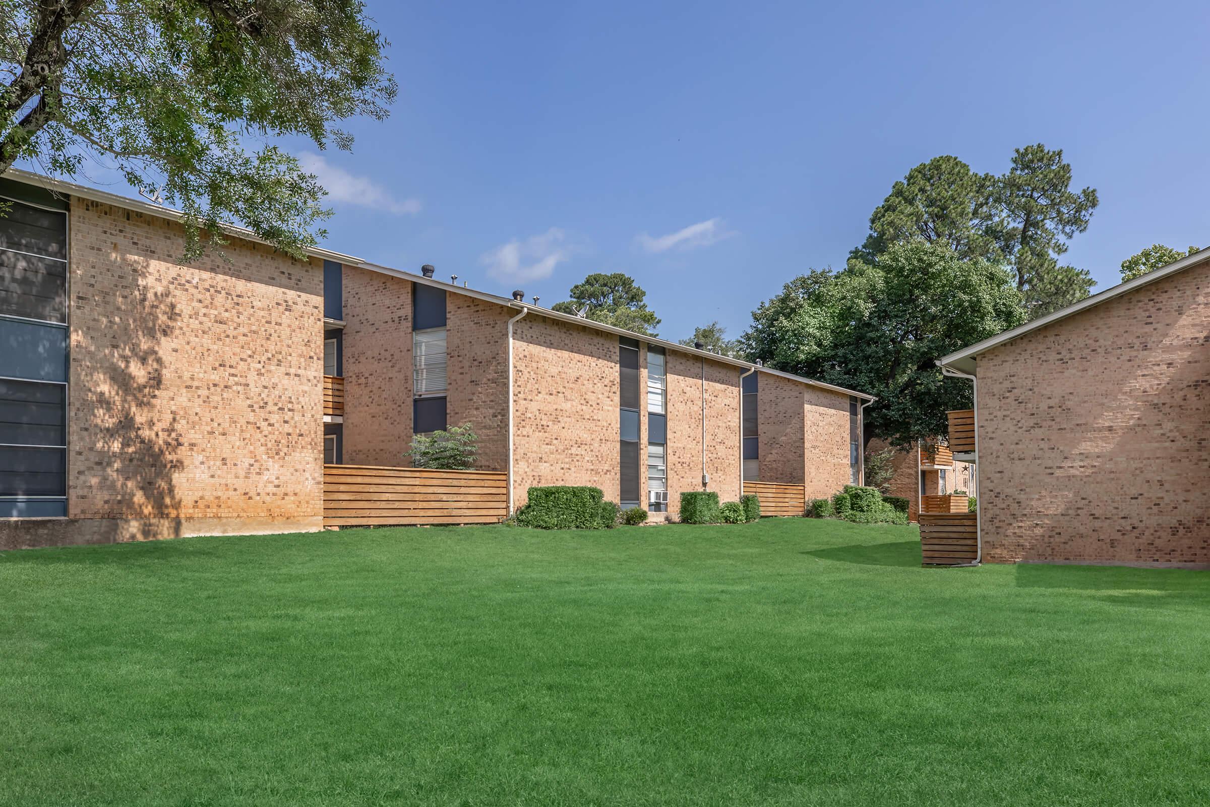 A view of two identical brick buildings surrounded by well-maintained green grass. The buildings feature large windows and wooden accents. Trees and shrubs are present, adding to the landscaped area. The sky is clear with a few clouds, indicating a pleasant day.