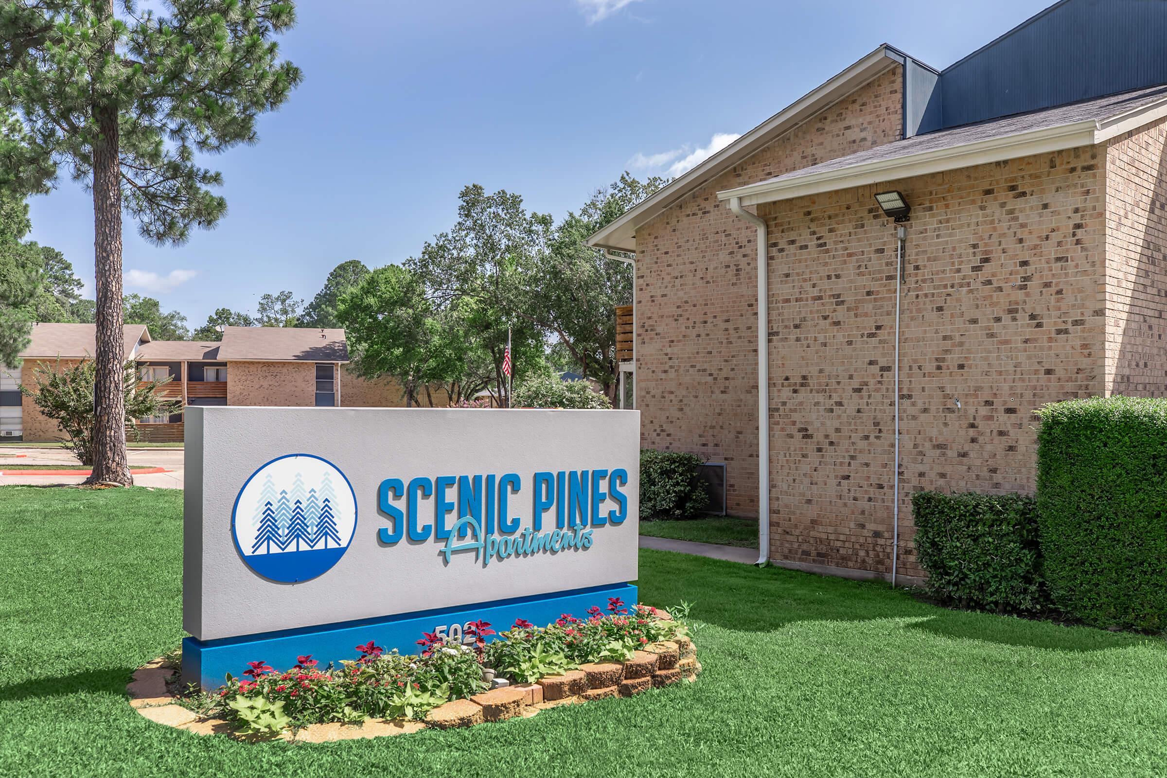 Sign for Scenic Pines Apartments, featuring blue and white colors with a graphic of pine trees. The sign is located in a well-maintained grassy area, surrounded by flower beds. In the background, there are residential buildings and trees, indicating a serene living environment.
