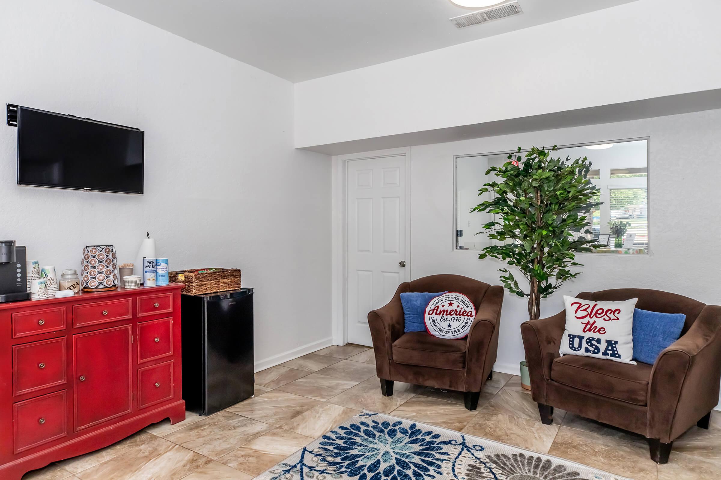 A cozy living space featuring a red cabinet, a mini fridge, and a flat-screen TV mounted on the wall. Two brown armchairs with decorative pillows reading "Bless the USA" and "America" are positioned around a blue floral rug. A potted plant adds a touch of greenery to the room.