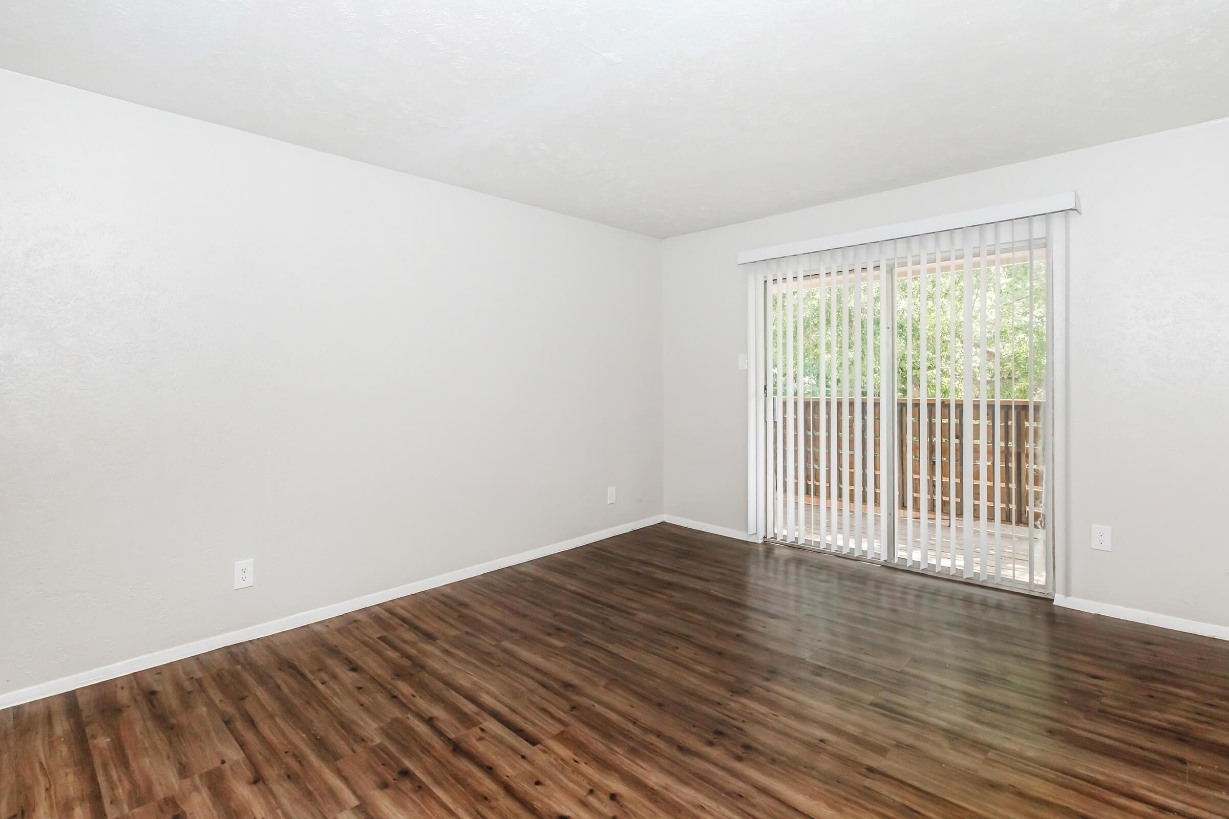 A spacious, empty room with light-colored walls and wood laminate flooring. It features large sliding glass doors with vertical blinds leading to a balcony, allowing natural light to enter the space. The overall atmosphere is bright and airy, suitable for various interior design options.
