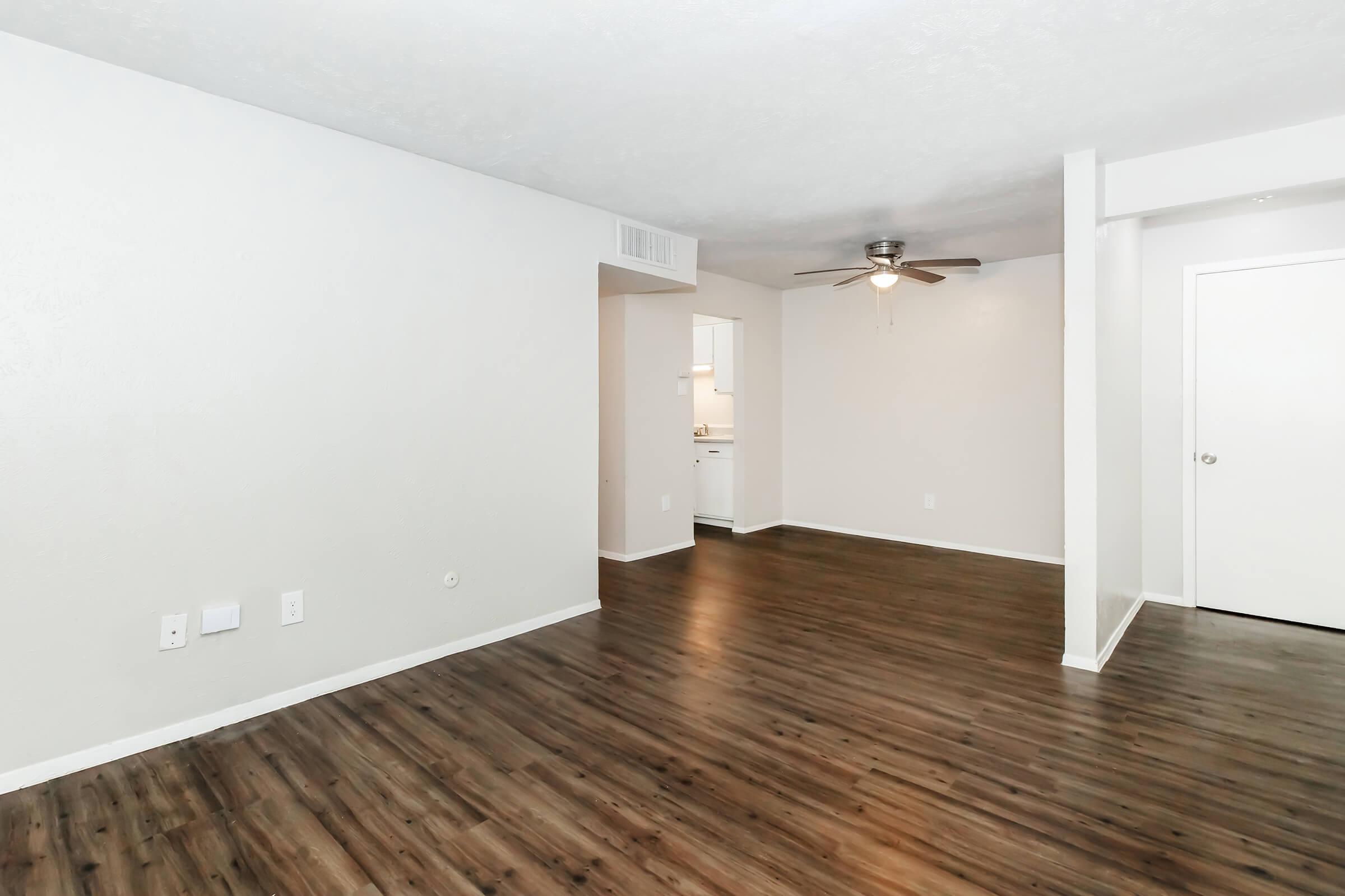 A spacious room with light gray walls and a brown laminate floor. There is a ceiling fan in the center, and a doorway leading to a kitchen area is visible in the background. The room is empty, creating a clean and open atmosphere.