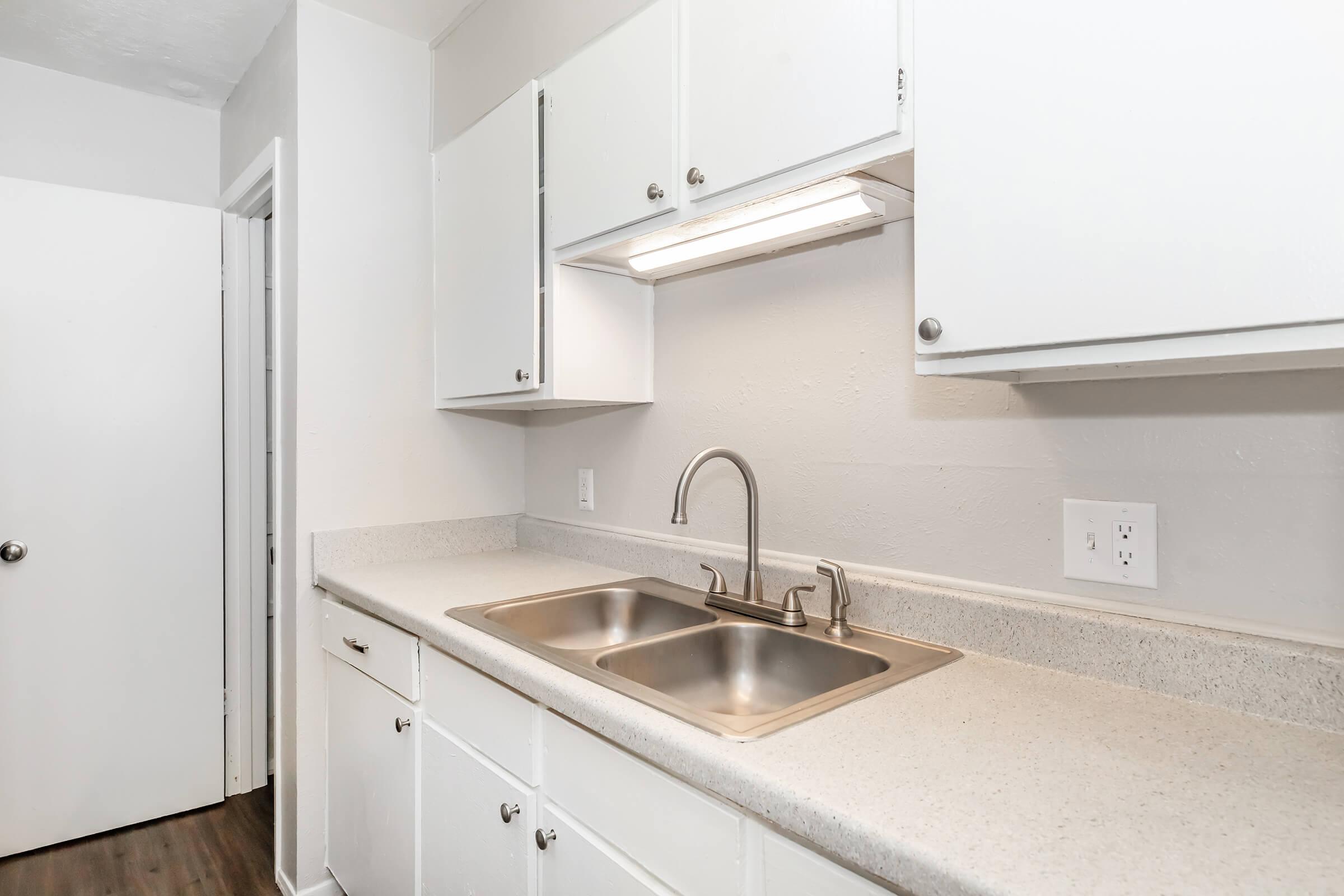 A modern kitchen featuring white cabinetry and a double sink. The countertop is light-colored, and there's an under-cabinet light illuminating the workspace. A door is visible in the background, leading to another room. The flooring is dark wood, adding contrast to the bright kitchen area.