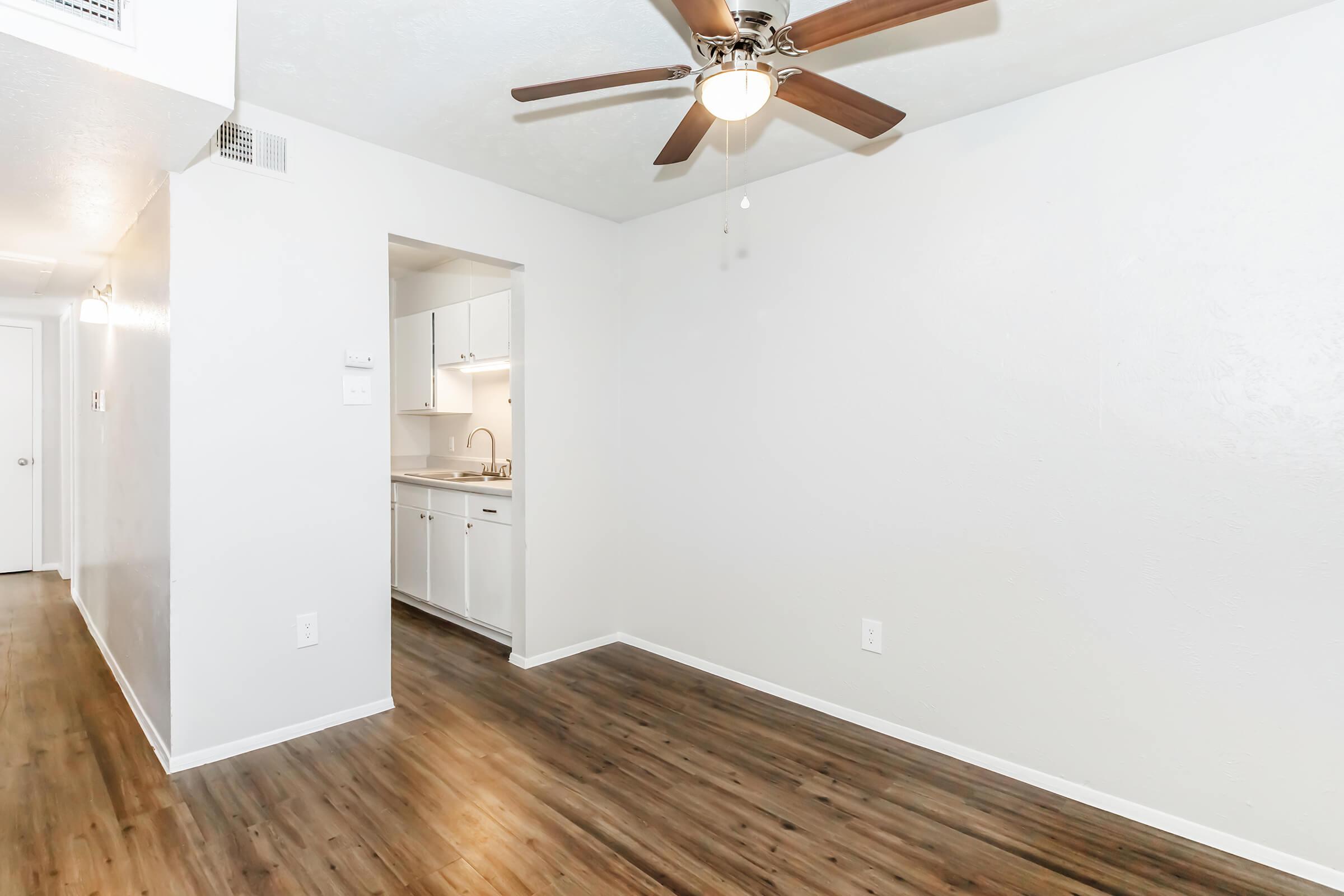 A bright and empty room with light gray walls and a wooden floor. A ceiling fan with wooden blades is visible. To the left, there is an open doorway leading to a kitchen area with white cabinets. The space is well-lit, creating an inviting atmosphere.