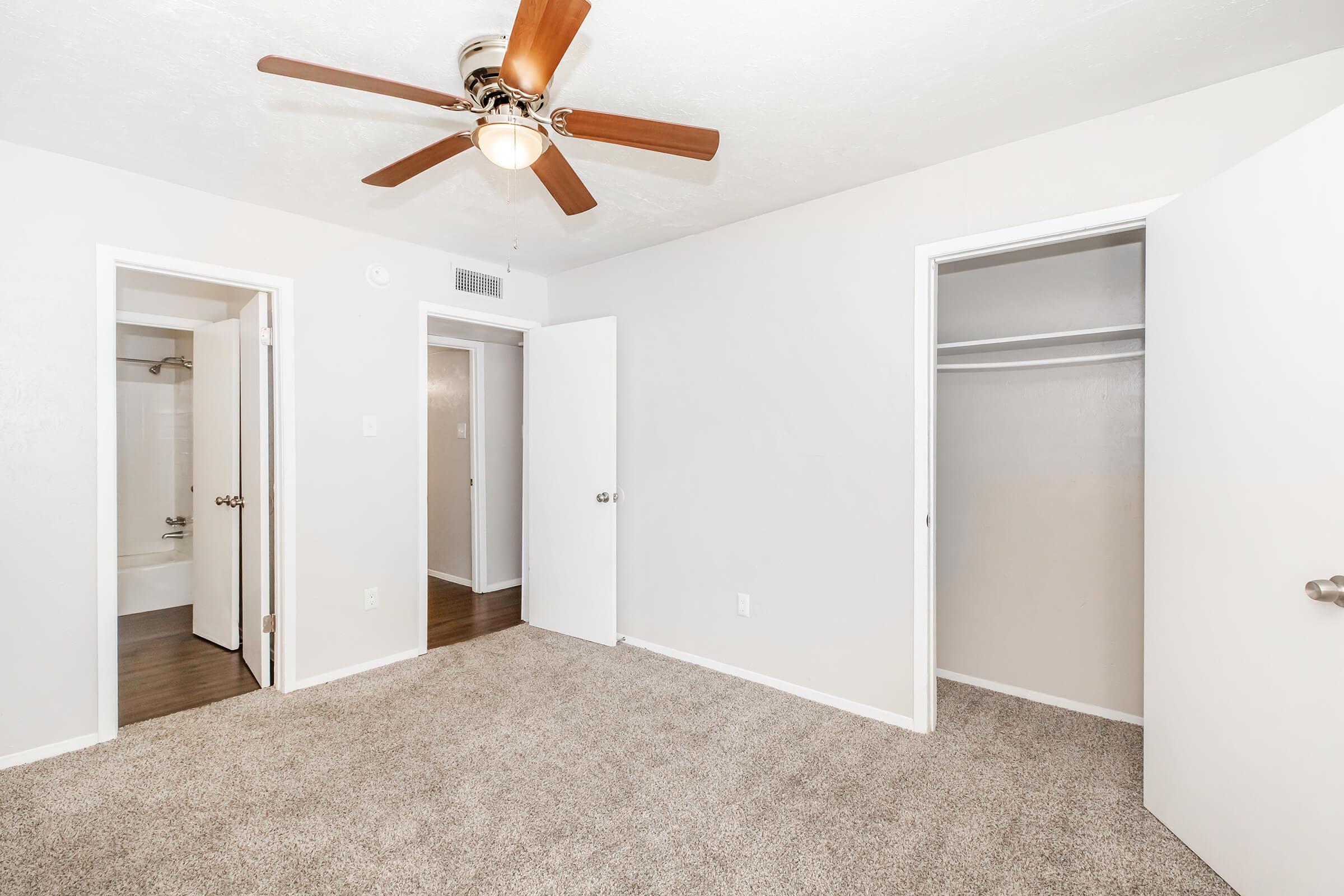 A well-lit bedroom featuring light gray walls and a ceiling fan. The room has two open doorways leading to additional spaces, and a large closet with sliding doors on the right. The floor is covered in plush beige carpeting, enhancing the cozy atmosphere.