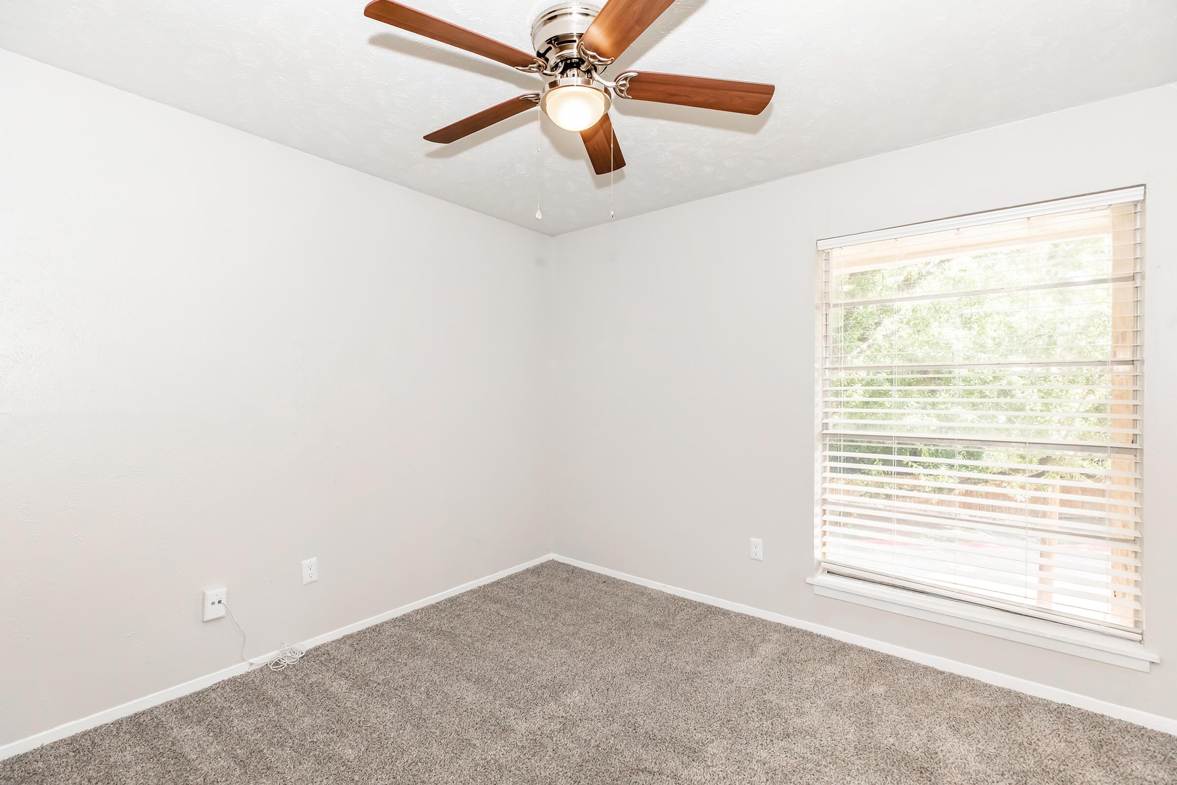 Empty room with light gray carpet, white walls, and a ceiling fan with wooden blades. A window with blinds allows natural light, showing greenery outside. The space appears clean and well-maintained, suitable for various uses such as a bedroom or office.